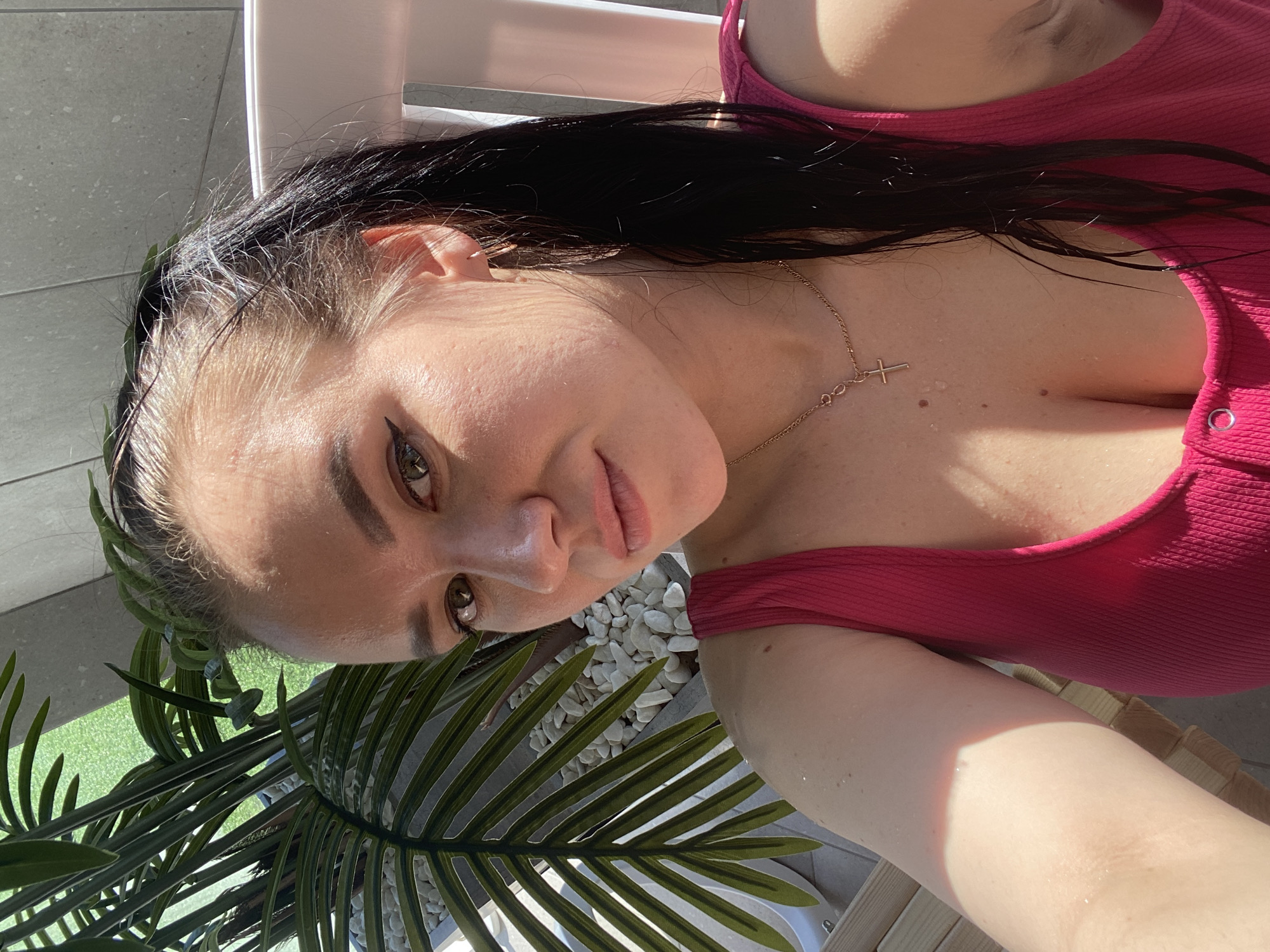 A selfie of a woman with dark hair pulled back, wearing a pink top and a cross necklace, is taken in an outdoor setting. Part of a palm-like plant is visible in the lower-left corner, along with white pebbles and a glimpse of green grass in the background. The location cannot be confirmed as Votkinsk, Russia, based solely on the visual information provided.