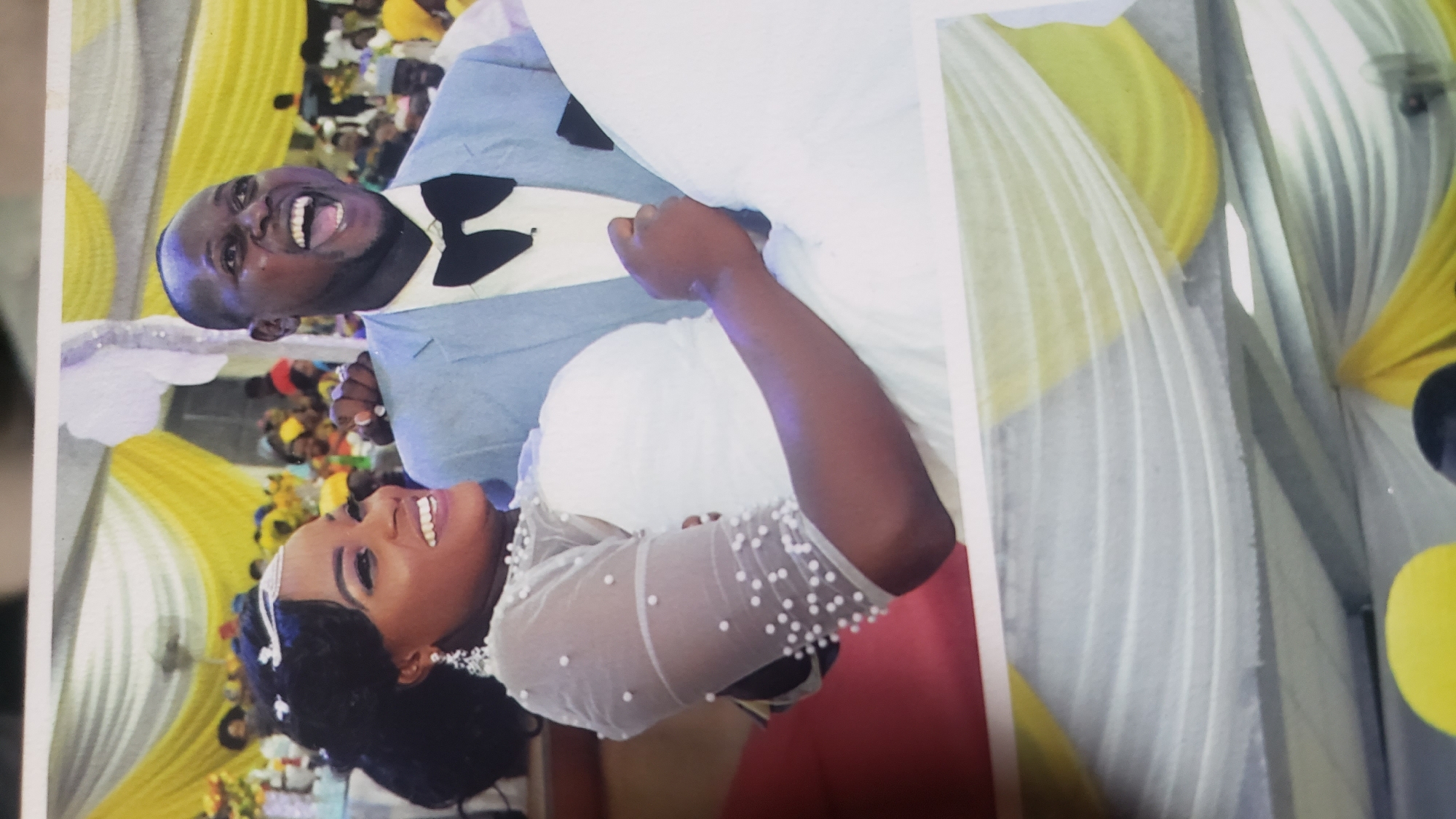 The media file shows a wedding celebration in Sabon Tasha, Nigeria. The primary subjects are a bride and groom in traditional wedding attire. The groom is wearing a light blue suit with a black bowtie and a white shirt. He is seen laughing heartily, holding the bride. The bride is wearing a white wedding dress adorned with pearls on the sleeves and a tiara. She is smiling radiantly, looking upwards. The background features yellow and white drapes, suggesting a decorated venue, likely a reception hall. In the blurred background, other wedding guests are visible, some wearing yellow and red attire. The overall atmosphere appears joyful and celebratory. The image captures a moment of happiness and connection between the newlywed couple.