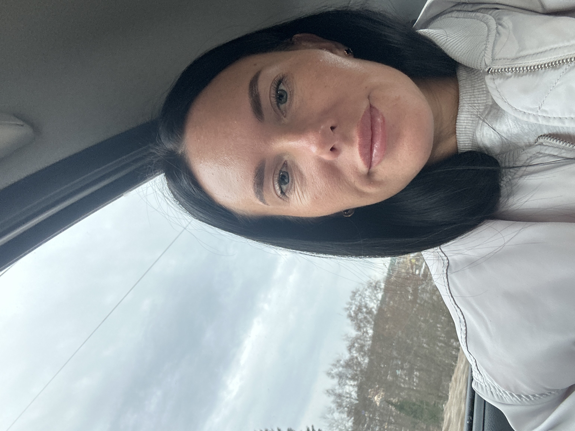 A young woman with dark hair and light eyes is taking a selfie in a car. She is wearing a light-colored jacket. The background shows trees and an overcast sky, possibly indicating a location in Ольховка, Russia. The image appears to be taken inside a vehicle, looking through the windshield, where there are trees and an overcast sky.