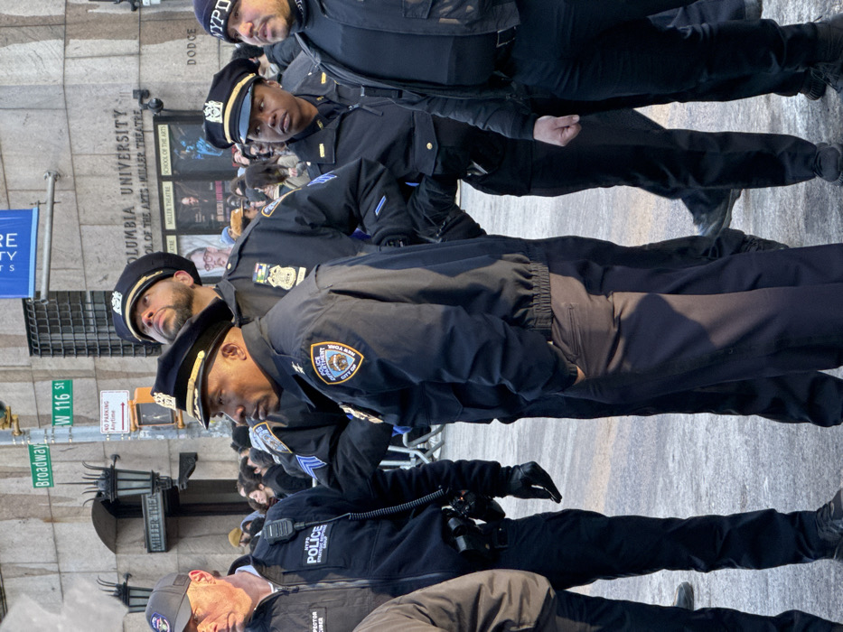 A group of NYPD officers stand in formation near Columbia University and Miller Theatre on Broadway at W 116 St. The officers wear dark blue uniforms and hats. One officer is labeled "Inspector Gruber." Signage indicates "No Parking Anytime." A sign reads "Columbia University School of the Arts Miller Theatre." A theater marquee is visible in the background.