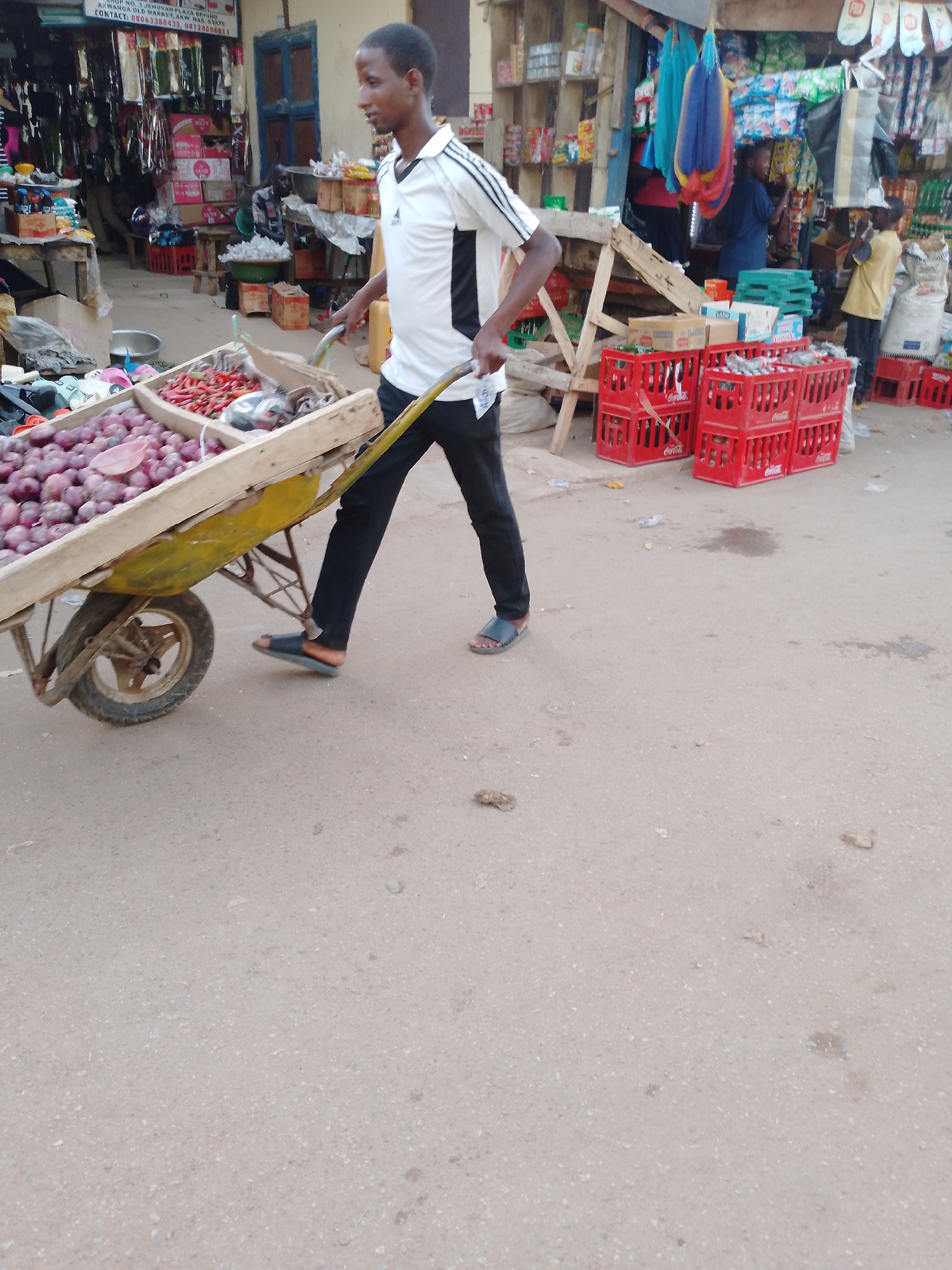 This daytime image depicts a bustling outdoor market scene in Akwanga, Nigeria. A young man, wearing a white and black short-sleeved polo shirt, black trousers, and sandals, is the central figure. He is walking from right to left, pushing a wooden wheelbarrow filled with produce. The wheelbarrow contains a large quantity of purple onions in the foreground and red chili peppers in another section.
In the background, market stalls line the area, displaying various goods such as packaged items, clothing, and household articles. Several red plastic Coca-Cola crates are stacked on the right side, suggesting a beverage vendor. Other people are visible in the background, tending to stalls or moving through the market. Overhead, colorful fabrics or hammocks are hanging, and a white banner with the word "FUN" is discernible. The ground is a light-colored, possibly unpaved or asphalt, surface.
A prominent sign in the upper left corner displays text: "NO. 3 JEHOVAN PLAZA BEHIND AKWANGA OLD MARKET, ANW. NAS STATE" and "CONTACT: 08063388433, 0812400681". The scene captures the everyday commerce and lively atmosphere of a local marketplace.