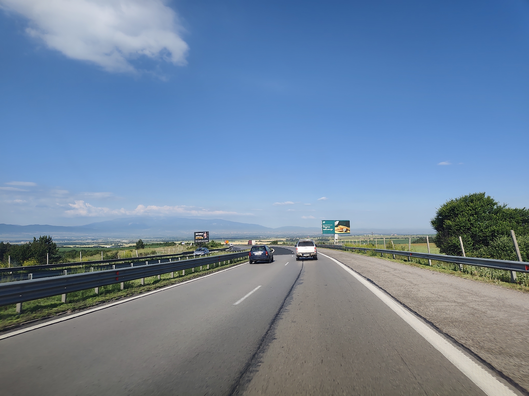 The image captures a highway scene on a clear day, with a blue sky and scattered clouds. The viewpoint is from inside a vehicle, looking forward along a multi-lane asphalt road. Two cars are visible in front, a dark blue hatchback and a white SUV. A metal guardrail runs along the right side of the road, separating it from a grassy verge and a line of trees and bushes beyond. In the distance, rolling hills and mountains form the horizon, with a hazy landscape of fields and structures in the valley. Several billboards are present along the roadside, one featuring a burger advertisement and another with a portrait and text. The scene is consistent with a highway outside a populated area, likely near the city of Eleshnitsa, Bulgaria, given the visual cues of the landscape and potentially the text on the billboards, although the text is not fully legible.