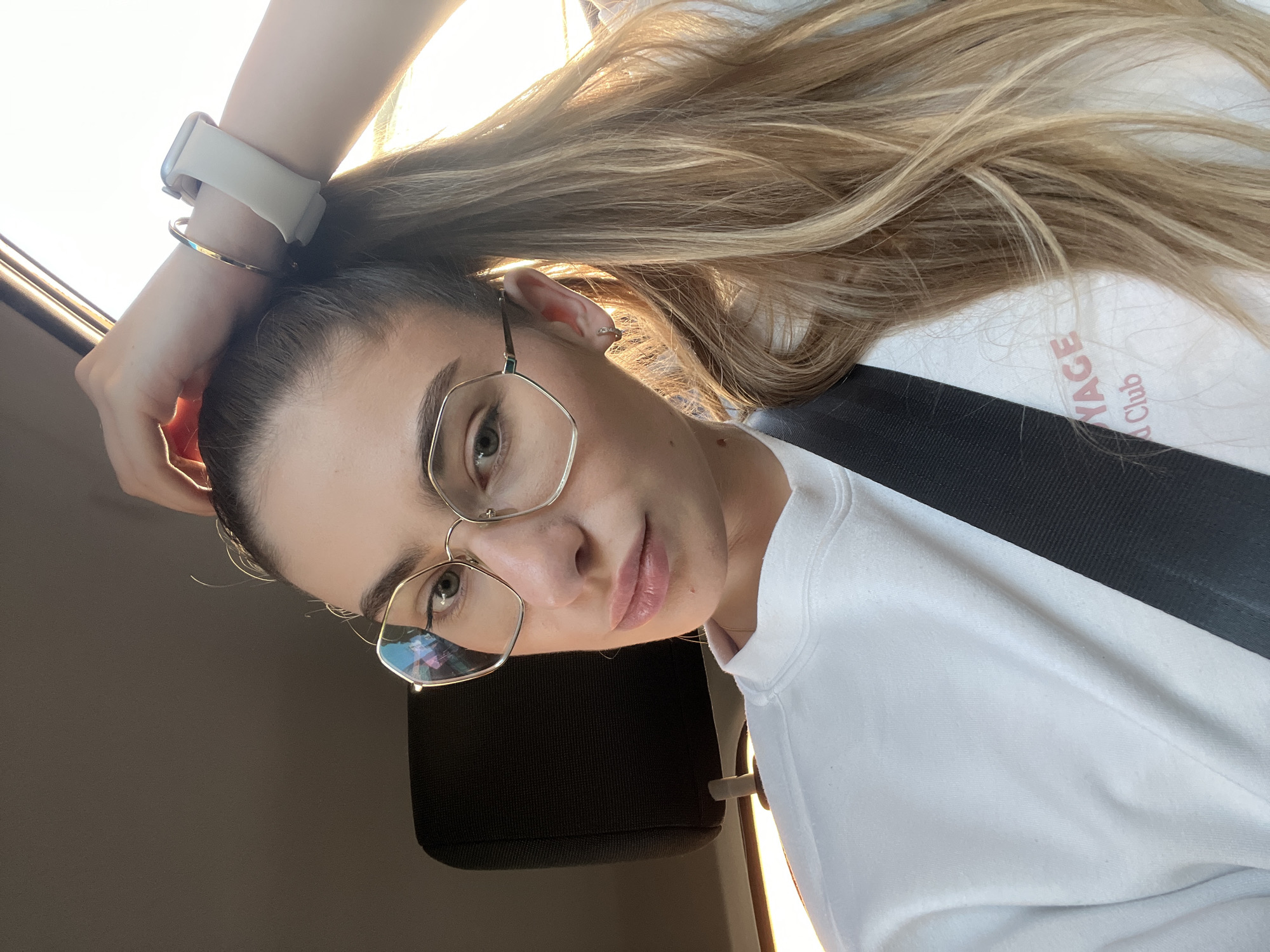 A woman with light brown hair, wearing glasses, a white sweater, and a seatbelt, is in a car. She is wearing a smart watch and a bracelet on her left wrist. The car's headrest is visible behind her. The location is Unknown, Unknown.