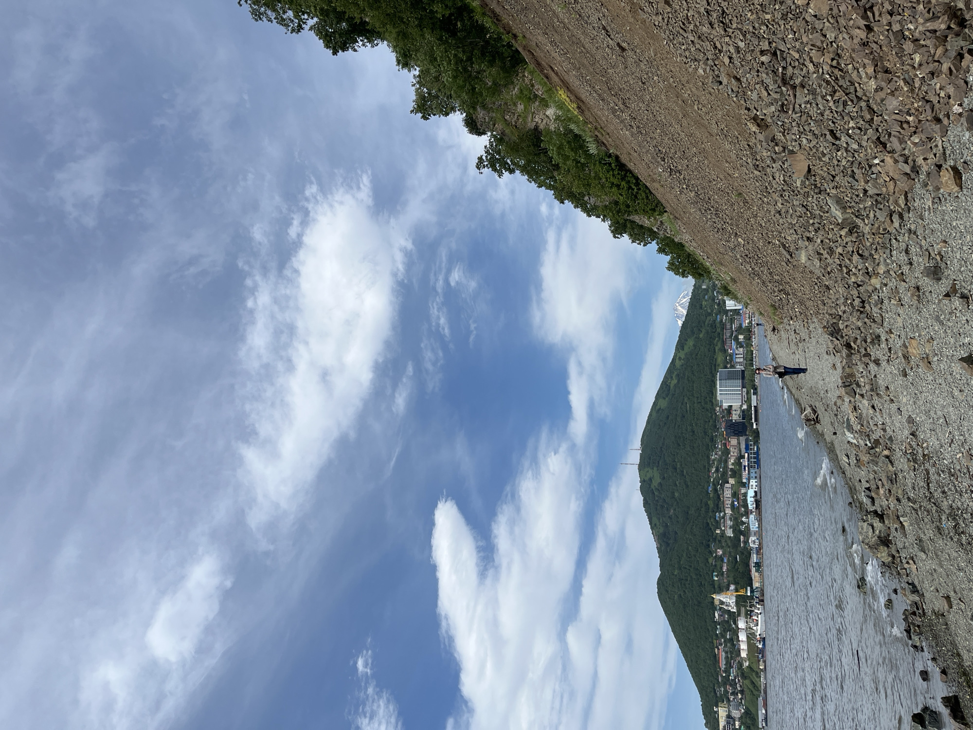 The image depicts a scene in Petropavlovsk-Kamchatsky, Russia, likely along the coastline. The foreground features a rocky shore with a person walking along it. The water is calm, with small waves lapping at the shore. In the mid-ground, a city stretches along the coast, with buildings of various sizes and architectural styles. A large, green hill dominates the background, topped with communication towers. To the right, a steep, rocky slope covered in trees descends towards the water. The sky is partly cloudy with wispy white clouds against a blue expanse. The overall impression is of a coastal city nestled against a natural landscape on a clear day.