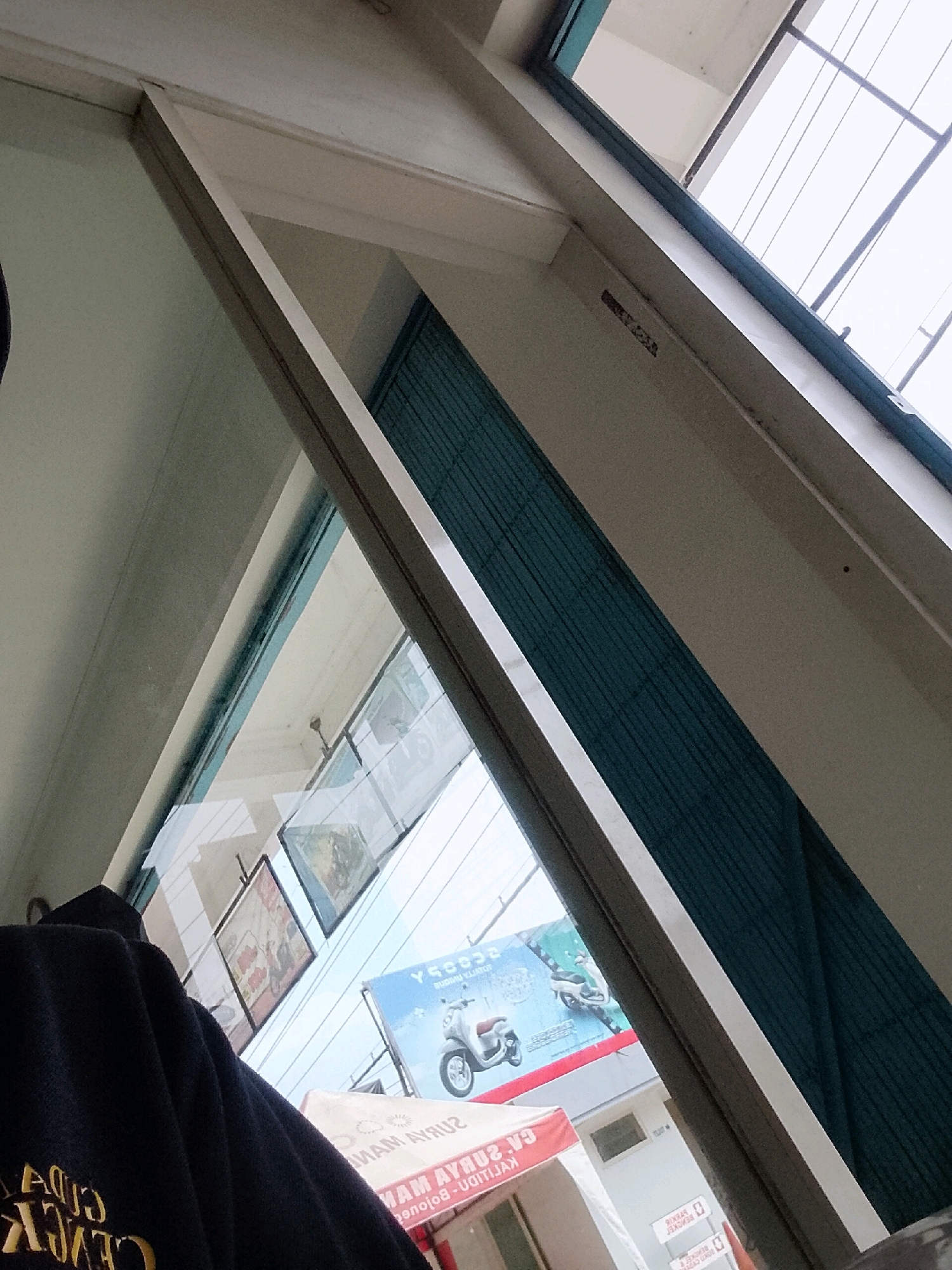 A person wearing a black hoodie is standing behind a large window, looking out at a street scene in Kalitidu, Indonesia. The hoodie has yellow lettering that reads "CITY CHICKEN". Outside the window, there are several billboards and signs, including one for a scooter. The architecture suggests a commercial area. The lighting is overcast, typical of a cloudy day in the region. The image is taken from a low angle, looking up towards the window and sky.