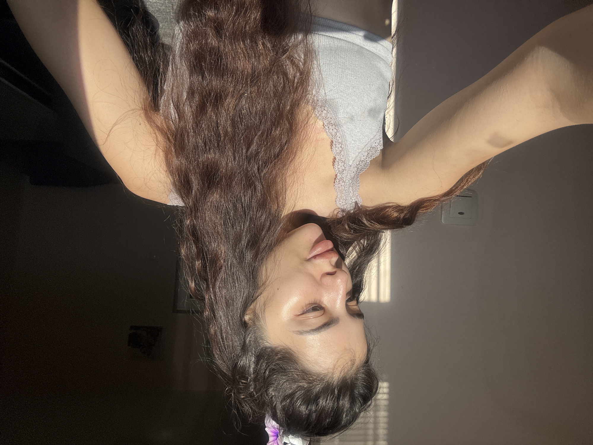 A young woman with long, wavy, dark brown hair is shown in an indoor selfie taken at an inverted angle. She is wearing a light gray tank top with lace detailing. Part of a flower is visible in her hair. The room is illuminated by sunlight streaming through blinds. Behind her, there are a wall, an electrical outlet, and blurry pictures hanging on the wall. The location is not identifiable as Plovdiv, Bulgaria from the given image.