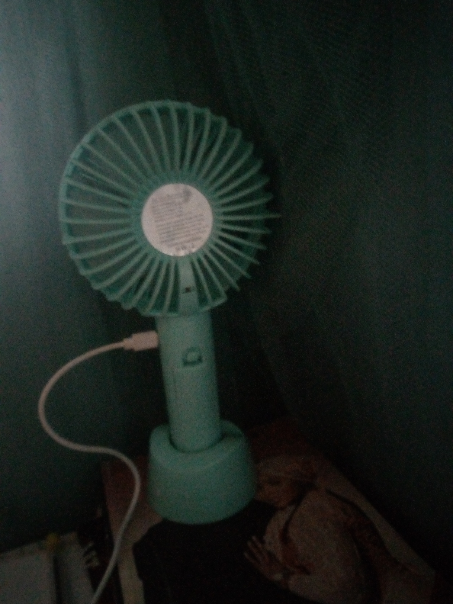 A teal, battery-powered fan with a white USB cord is positioned on a dark surface in front of a dark green, textured background. The fan is standing upright with its base on a book. The fan blades are fanned out and a circular label is visible in the center of the fan. A USB cable is connected to the fan's handle, with the other end extending out of frame. A person's chest and arm, wearing a patterned garment, are visible in the lower right corner of the image, suggesting the fan might be in a personal space. The scene appears to be indoors, possibly in a bedroom or living area, in Unguwan Rimi, Nigeria.