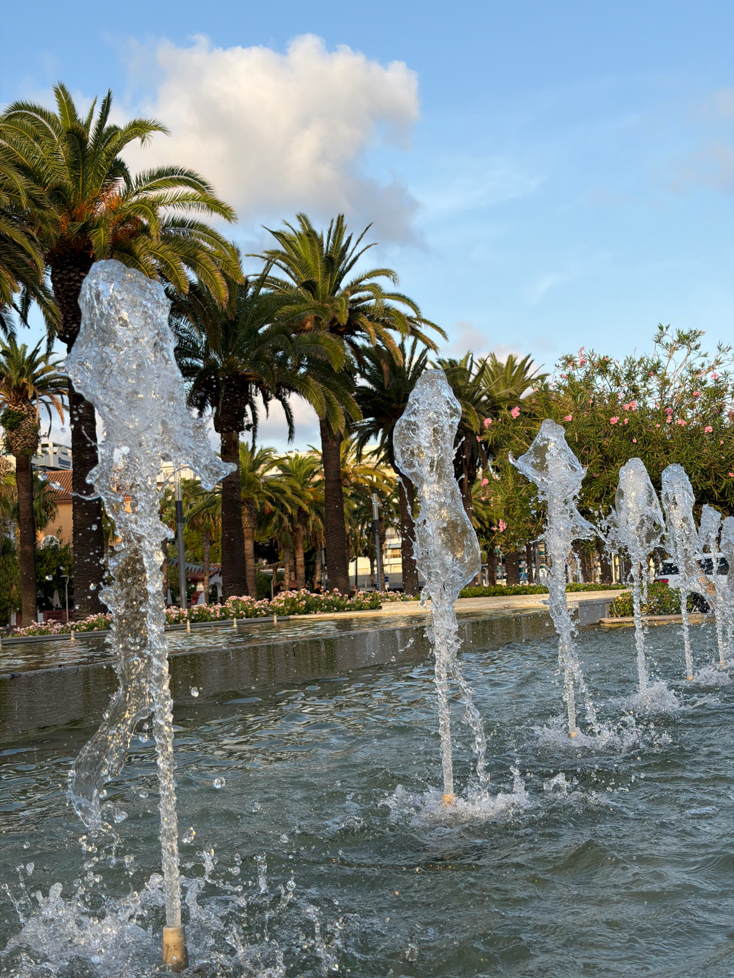 This is a low-angle, close-up photograph of a water fountain with multiple jets of water shooting upwards. The water jets are in the foreground, with the closest one appearing large and translucent, refracting light. Behind the fountain, a row of palm trees lines a pathway or promenade. In the background, there are buildings and more trees, some with pink flowers. The sky is blue with scattered white clouds. This scene is located in Salou, Spain, specifically on the Jaume I promenade, a popular tourist area known for its fountains and palm trees. There are no discernible people or specific actions captured in the frame. The environment suggests a warm, sunny day.