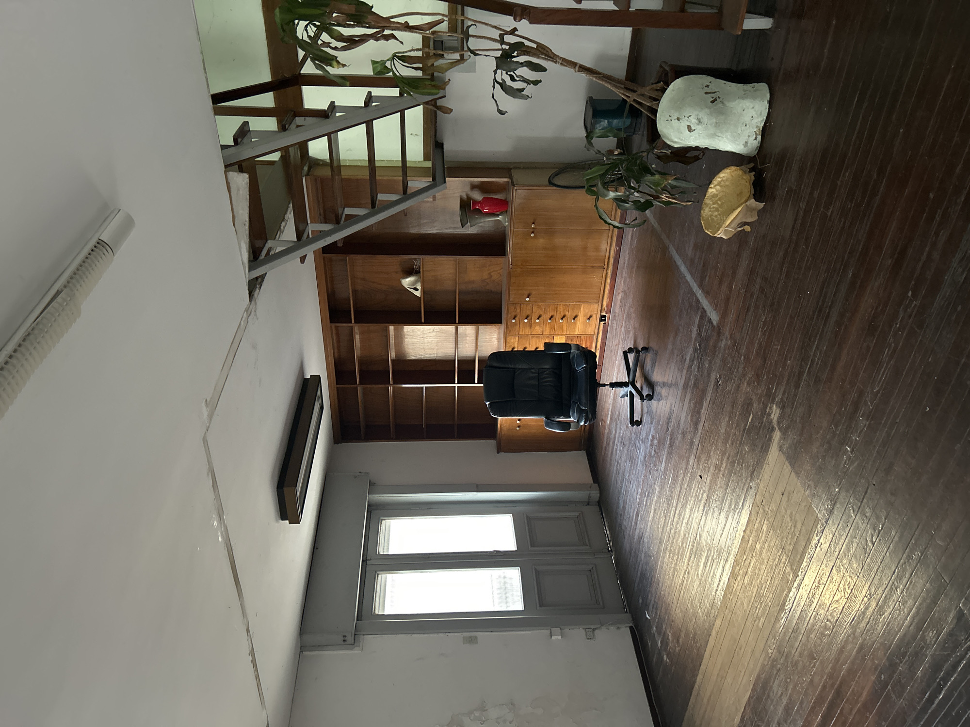 The image captures the interior of a room, possibly a former office or study, in a state of disuse or transition. The room is characterized by hardwood flooring and a large wooden bookcase spanning nearly the entire height of one wall. In front of the bookcase, a black office chair is positioned on the wooden floor. Natural light enters through a set of double doors at the far end of the room, casting bright reflections on the floorboards. Various objects are scattered around, including a tall plant, a pale-colored sculpture, and what appears to be a mushroom-shaped object. A metal staircase is visible leading upwards, suggesting a multi-story building. A dark rectangular air conditioning unit is mounted high on the wall, and a long, white fluorescent light fixture is also on the ceiling. The walls are white, with signs of wear and tear. The architectural style and the presence of hardwood floors suggest an older building, possibly located in a historic neighborhood of Buenos Aires, Argentina, though specific identifying landmarks are absent from the image.