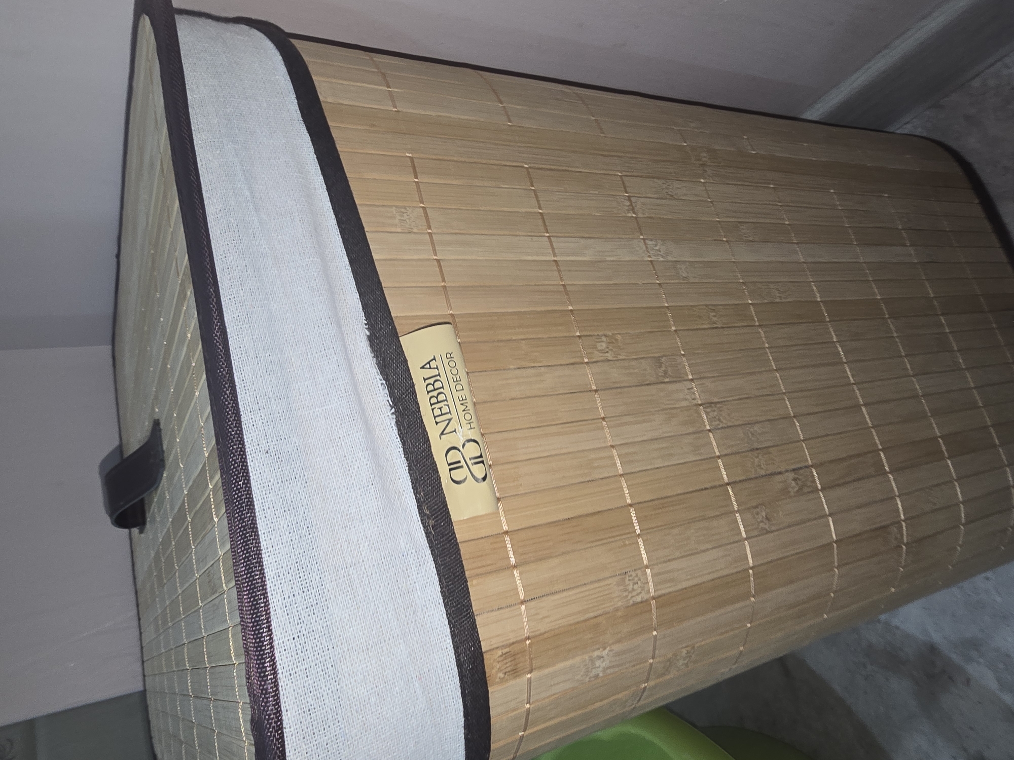 A light-colored bamboo laundry hamper with a dark trim and a dark brown handle on the lid sits in a dimly lit room. A beige label on the side of the hamper reads "NEBBIA HOME DECOR". The hamper is tapered and appears to be empty. To the left of the hamper, a portion of a bright green plastic object is visible. The background is a pale pink wall and a grey floor. The scene does not provide information about the city of Ulan-Ude, Russia.