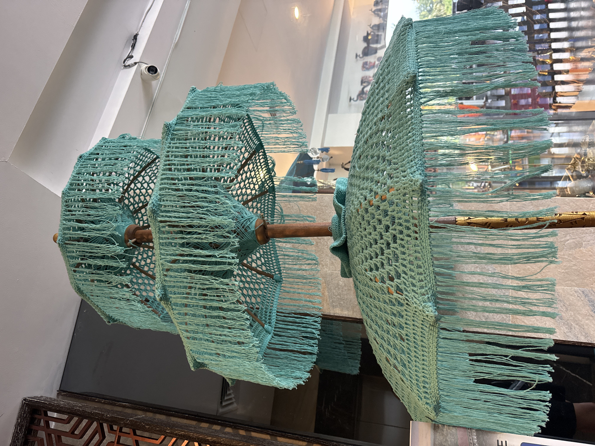 The image shows two decorative objects resembling fringed umbrellas suspended from a wooden rod, likely in an interior space in Pecatu, Indonesia. The "umbrellas" are constructed with turquoise-colored netting and long fringe. A security camera is visible mounted on the wall above the left-most umbrella. A blurred window shows an assortment of items and a person reflected in it. The objects are placed above a dark-colored, patterned surface.