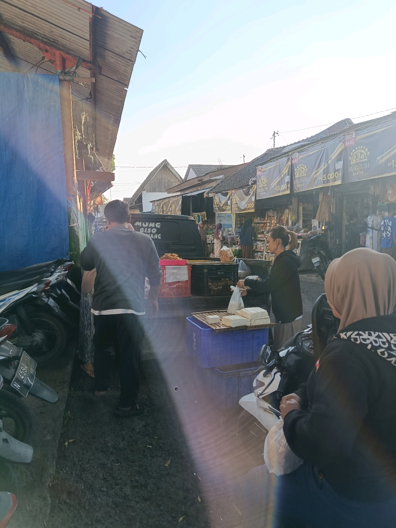 The image depicts an outdoor market scene in Bululawang, Indonesia. In the foreground, two individuals are visible. A person wearing a black hooded jacket and a headscarf is seated on a motorcycle, holding a plastic bag. Beside them, another person, partially obscured, wears a dark hooded jacket and also appears to be holding a plastic bag.
The middle ground reveals a woman actively engaged in a transaction, holding a plastic bag and interacting with a stall. In front of her, on a wooden structure, are several blocks of tofu. Behind her, a dark colored van with text that includes "MUNG BISO NANG" is parked. Red plastic crates containing goods are also present. To the left, motorcycles are parked along a structure covered with a large blue tarp.
The background shows several market stalls with awnings, some displaying advertisements for "MAGNUM KRETEK" with prices listed as "RP 15.000". Other people are seen browsing or working at the stalls. The overall environment suggests a bustling local market with a clear sky overhead and sunlight casting shadows. The ground appears to be paved with some debris, including fallen leaves.