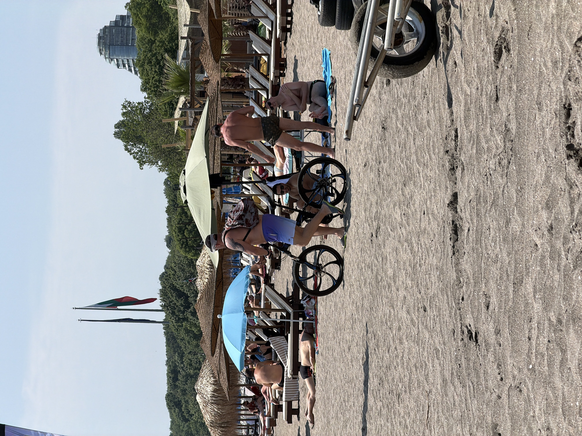 This image depicts a sunny beach scene in Burgas, Bulgaria, specifically identifiable by the distant Grand Hotel Millennium Burgas and the Bulgarian flag visible on a flagpole. The foreground features dark, fine-grained sand with numerous footprints and ripples.
In the mid-ground, a man with visible tattoos on his left arm, wearing a backpack, light blue ombre swim shorts, and flip-flops, walks from left to right while pushing a black bicycle with large, spoked wheels. To his left, another man lies face down on the sand beneath a light blue fabric umbrella. Directly behind them, multiple individuals relax on beige sun loungers under a row of straw-thatched umbrellas.
Further to the right, a man with his back to the viewer stands near a large olive-green fabric umbrella, wearing dark patterned swim trunks. To his right, another man sits on a blue towel on the sand, wearing dark swim trunks and sunglasses, holding a small object in his hands. A partial view of a metal cart with a single wheel and stacked tires is visible in the bottom right corner.
In the background, a dense line of green trees and bushes separates the beach from a distant cityscape. Two tall flagpoles stand on a hill above the treeline; one displays the Bulgarian flag (white, green, red horizontal stripes), while the other holds a dark-colored flag. To the right of the flagpoles, the distinctive, multi-tiered, glass-fronted building of the Grand Hotel Millennium Burgas rises above the trees. The sky is clear and light blue with minimal cloud cover.