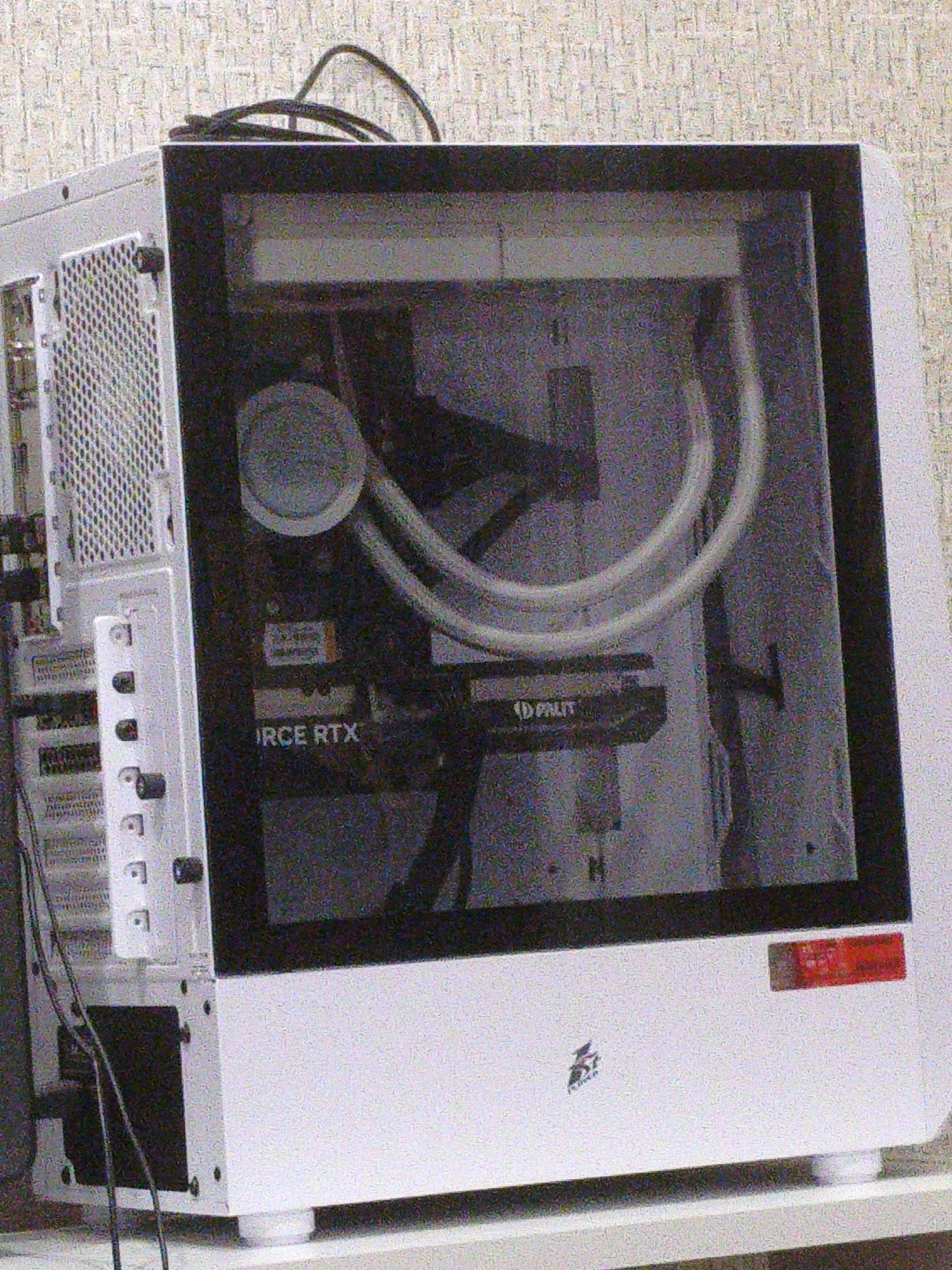 The image shows a white computer tower with a glass side panel, revealing internal components. The computer is on a light-colored surface, possibly a desk or shelf, in front of a textured, light-colored wall. Visible internal components include a graphics card with "IRCE RTX" printed on it, a circular water cooler pump, and tubing for a liquid cooling system. The computer appears to be in an indoor environment. There is no indication of the location being Chelyabinsk, Russia.