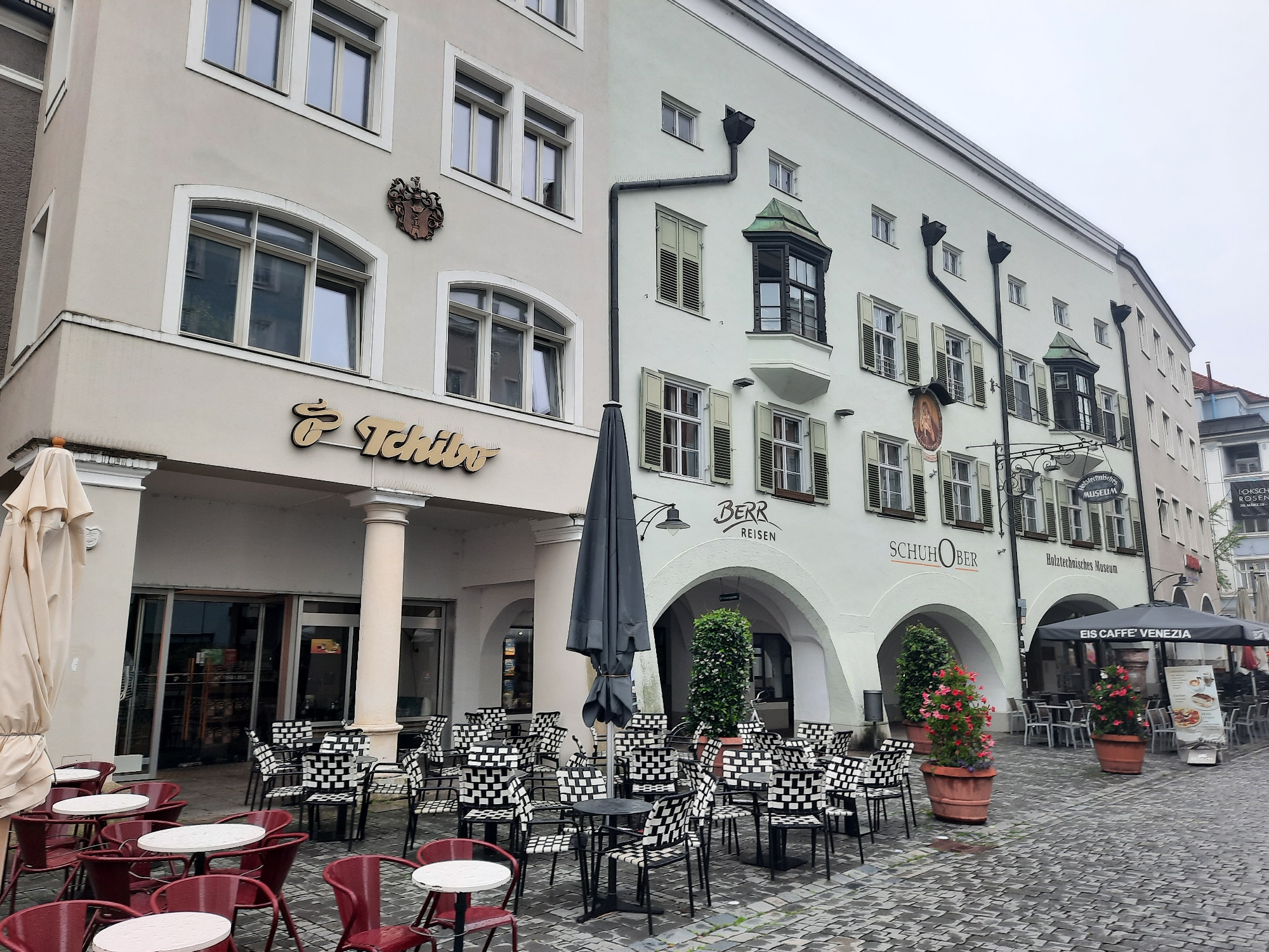 The image depicts a street scene in Rosenheim, Germany, likely during overcast weather. The main focus is on the facade of a building housing several businesses. In the foreground, an outdoor seating area for a cafe is set up with numerous tables and chairs. Red chairs surround white-topped tables on the left side of the image, while black and white checkered chairs surround black tables on the right. A large, closed black umbrella stands prominently in the center, suggesting it is not currently in use due to the weather or time of day.
The building's facade features a cream-colored section on the left with arched windows and a prominent "Tchibo" logo. To the right, the building's facade is a lighter, greenish hue with arched entrances. Signs for "Berr Reisen," "Schuh Ober," "Holztechnisches Museum," and "EIS CAFFÈ VENEZIA" are visible, indicating the businesses located within. Two potted plants with red and pink flowers are placed in front of one of the arched entrances. The ground is paved with cobblestones, which appear wet, further suggesting recent rain. In the background, other buildings and parts of the city are visible. There are no people actively depicted in the scene, though the setup implies a place of commerce and public access.