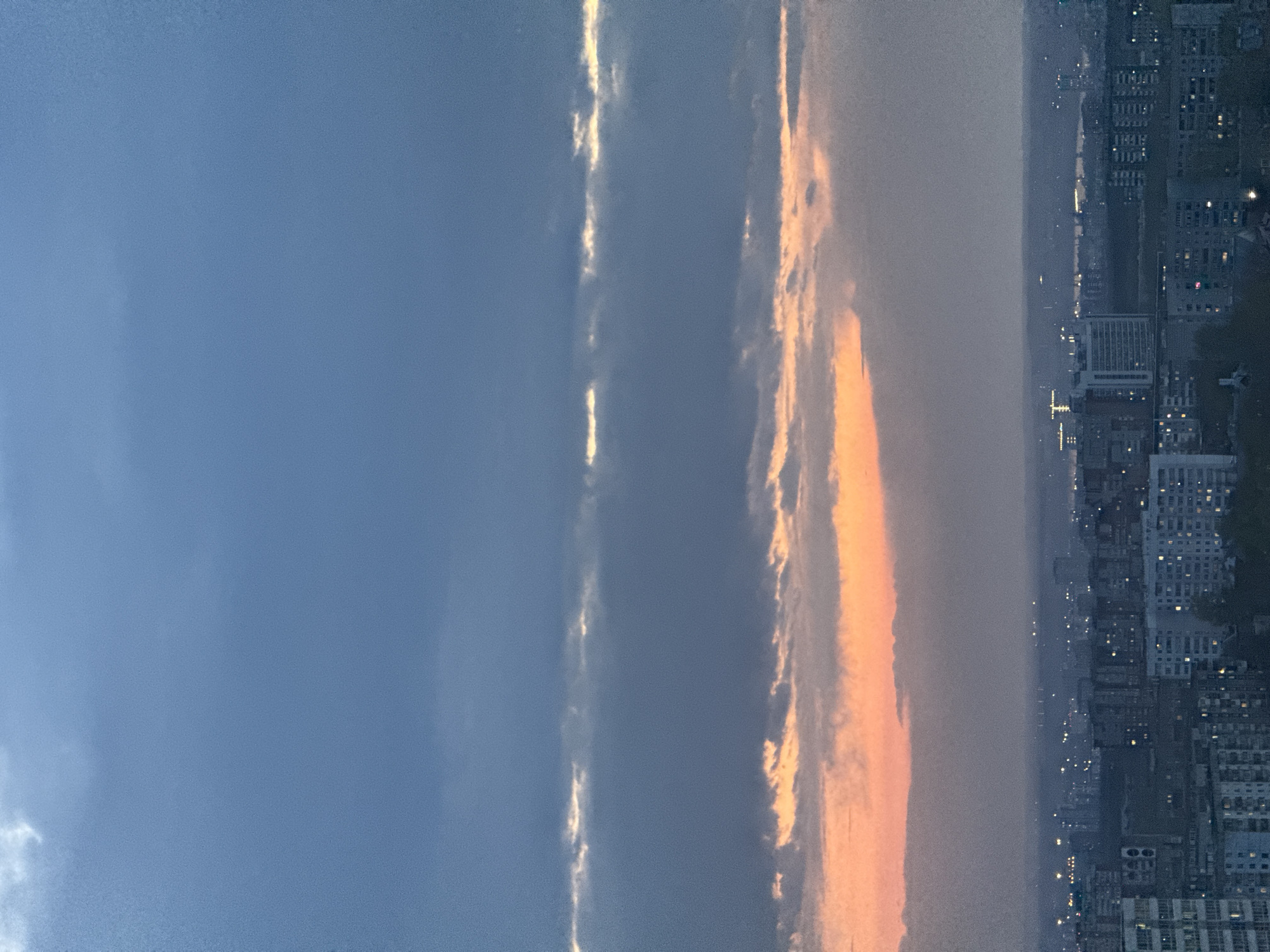 The image shows a twilight view of what appears to be a cityscape, possibly Perm, Russia, due to the query. The buildings are densely packed and lit with lights, suggesting it is night. The sky is a mix of dark blue and orange hues, indicating either sunset or sunrise. There are also distinct clouds.