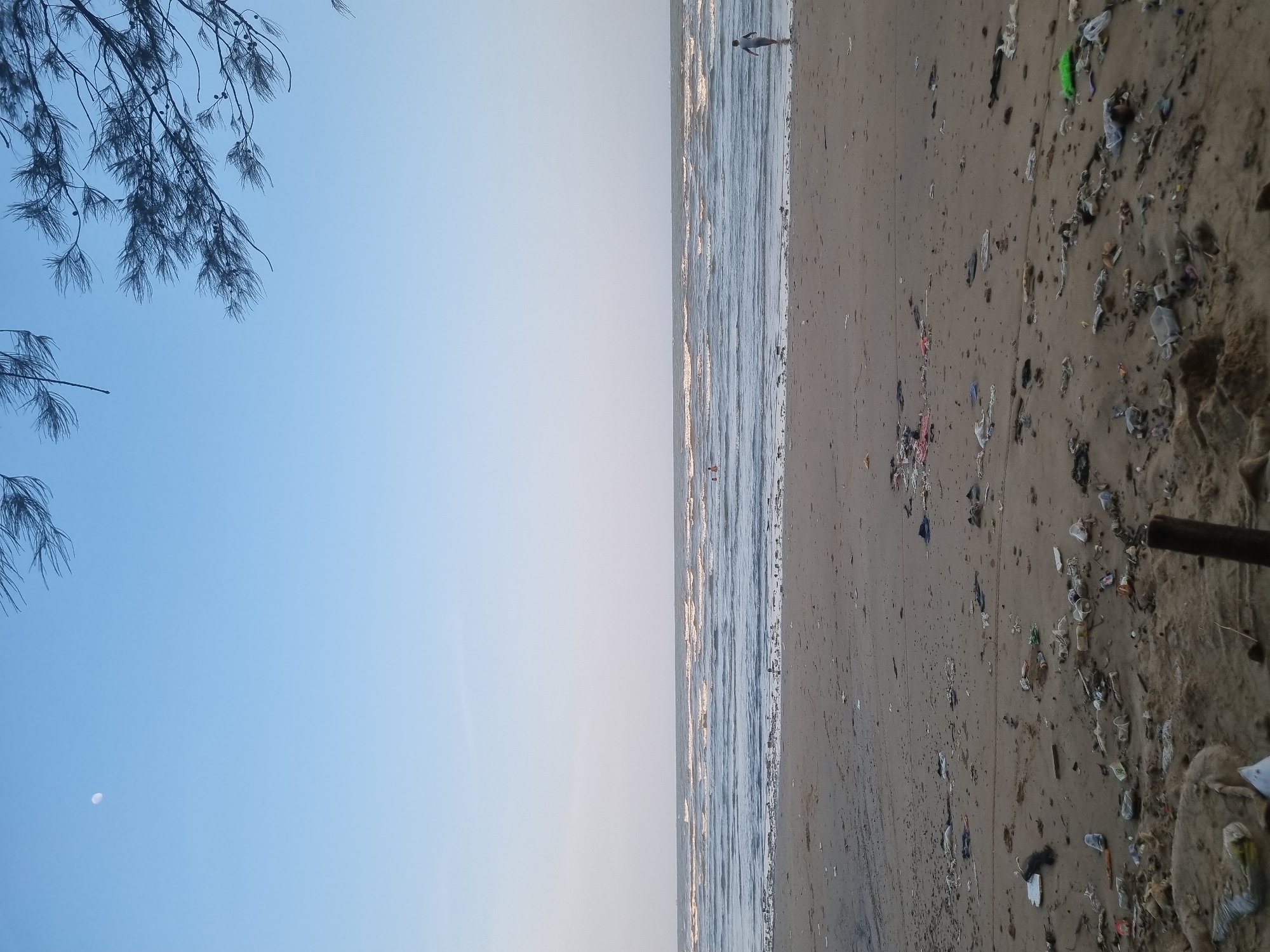 This image depicts a beach scene in Balikpapan, Indonesia, characterized by a vast expanse of sand littered with debris. The ocean stretches to the horizon, with gentle waves rolling towards the shore. Several individuals are visible in the water, some appearing to be swimming or wading. A lone person stands at the edge of the water, facing away from the camera. The sand itself is heavily polluted with a variety of trash, including plastic bottles, wrappers, and other discarded items, suggesting a significant environmental issue. Overhead, tree branches frame the top of the image against a clear, pale blue sky, where a small crescent moon is visible. The overall impression is one of a natural landscape marred by human-generated waste.