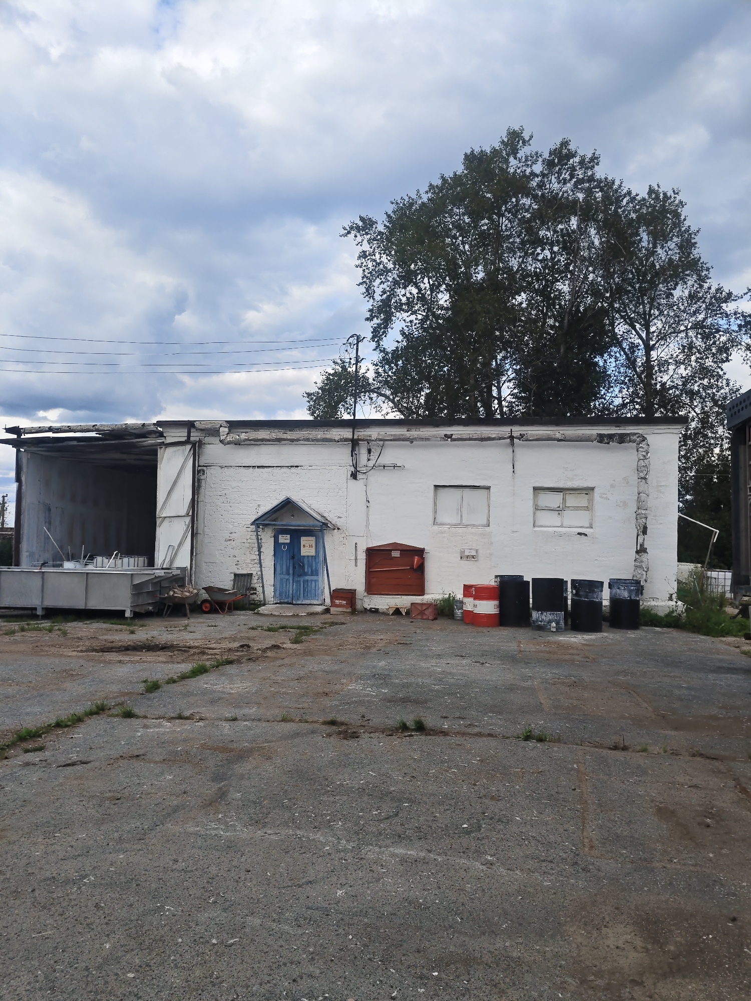 The image depicts an outdoor industrial or storage area in the city of Platoschino, Russia. A long, white, brick building with a black roof dominates the scene. The building has a large, open bay on the left side, revealing some white shelving or containers inside. To the right of the open bay is a small blue door with a triangular roof overhang. A sign with "8-16" is visible on the door. There are two windows on the right side of the building, both with white-paned glass. In front of the building, a collection of industrial barrels, some red and white, and others black, are arranged on the ground. A red metal container is also present. To the left of the building, near the entrance, there is a wheelbarrow with some wood. The ground is a cracked asphalt or concrete surface with patches of dirt and grass growing through. In the background, several mature trees with dense foliage are visible against a cloudy sky. Power lines and a utility pole are also present in the upper portion of the image. The overall impression is one of a functional, somewhat neglected, working area.