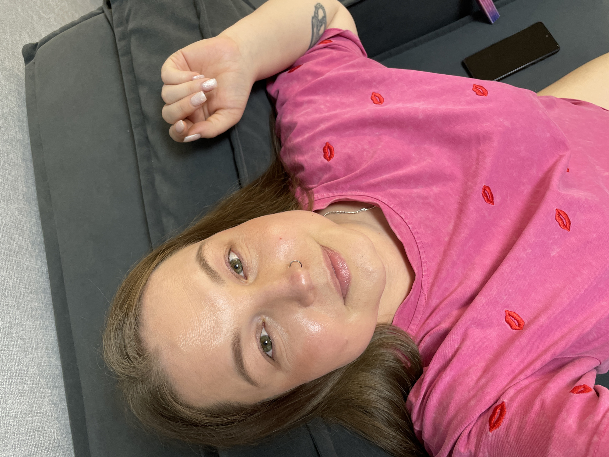 An overhead, eye-level photograph taken in Pervomaysky, Russia, shows an adult female lying supine on a dark grey upholstered sofa. She has medium-length brown hair, green eyes, and light makeup. A silver hoop nose piercing is visible in her left nostril. She is wearing a hot pink t-shirt with multiple embroidered red lip designs and a silver chain necklace. Her left hand, with light, shimmery nail polish, rests near her head. A dark-colored tattoo is partially visible on her right upper arm. To her right, on the sofa, a black smartphone and a small, partially obscured purple object (possibly a nail file) are present. The background behind the sofa features a light grey textured wall. The individual is looking directly at the camera with a subtle smile.