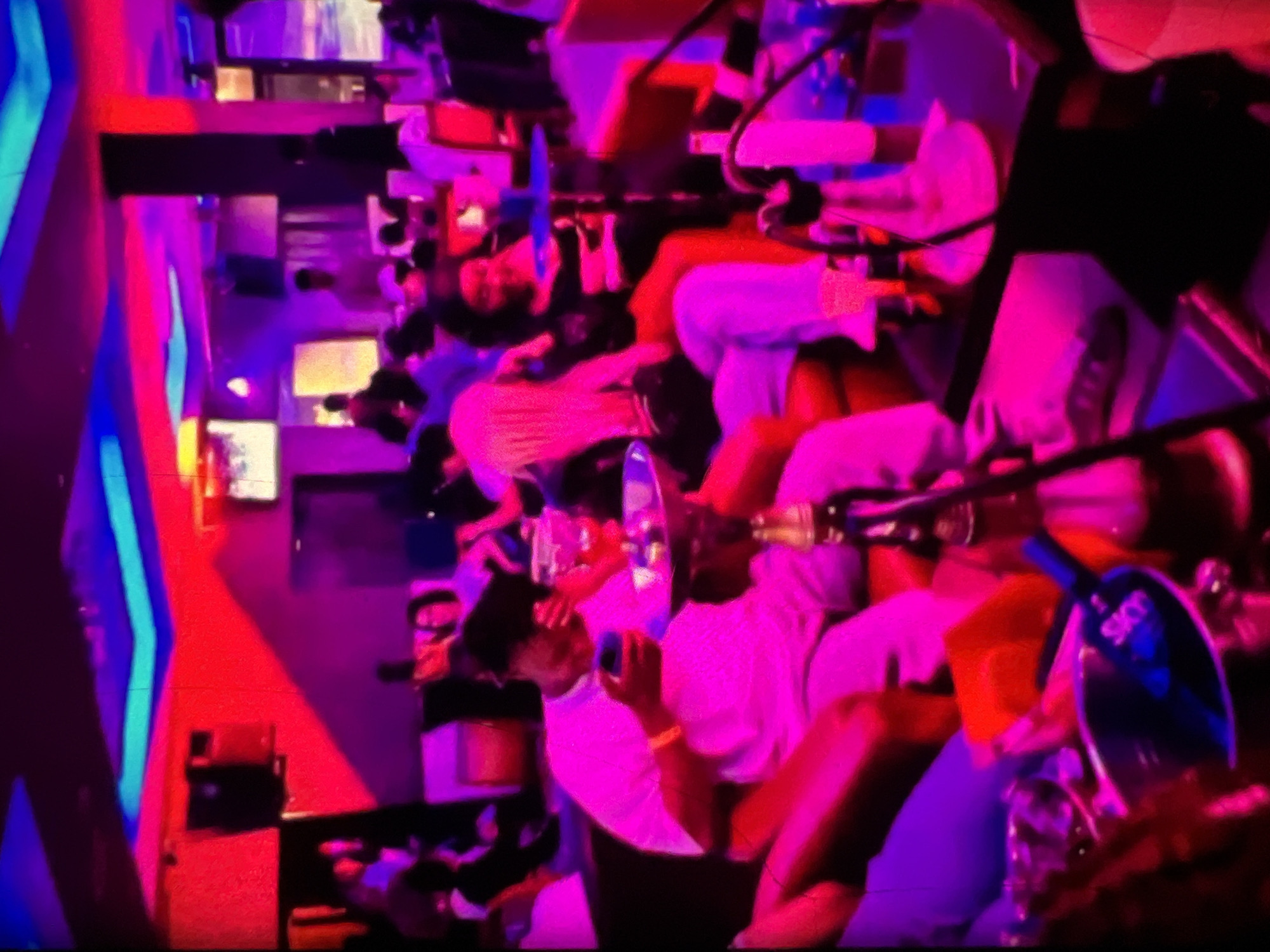 The image depicts a dimly lit, vibrant nightclub or lounge environment, likely in Lagos, Nigeria, characterized by neon blue and magenta lighting. Several individuals are present, seated and engaged in various activities. In the foreground, a man in a white t-shirt is seated, holding a smartphone. To his right, a hookah pipe is prominently displayed on a table, with smoke or vapor visible around it. Further back, a woman with dark curly hair is seated and smiling, facing towards the right of the frame. Other patrons are visible in the background, some appearing to be interacting, while others are focused on their own activities. The overall atmosphere suggests a social gathering with music and entertainment, common in Lagos nightlife.