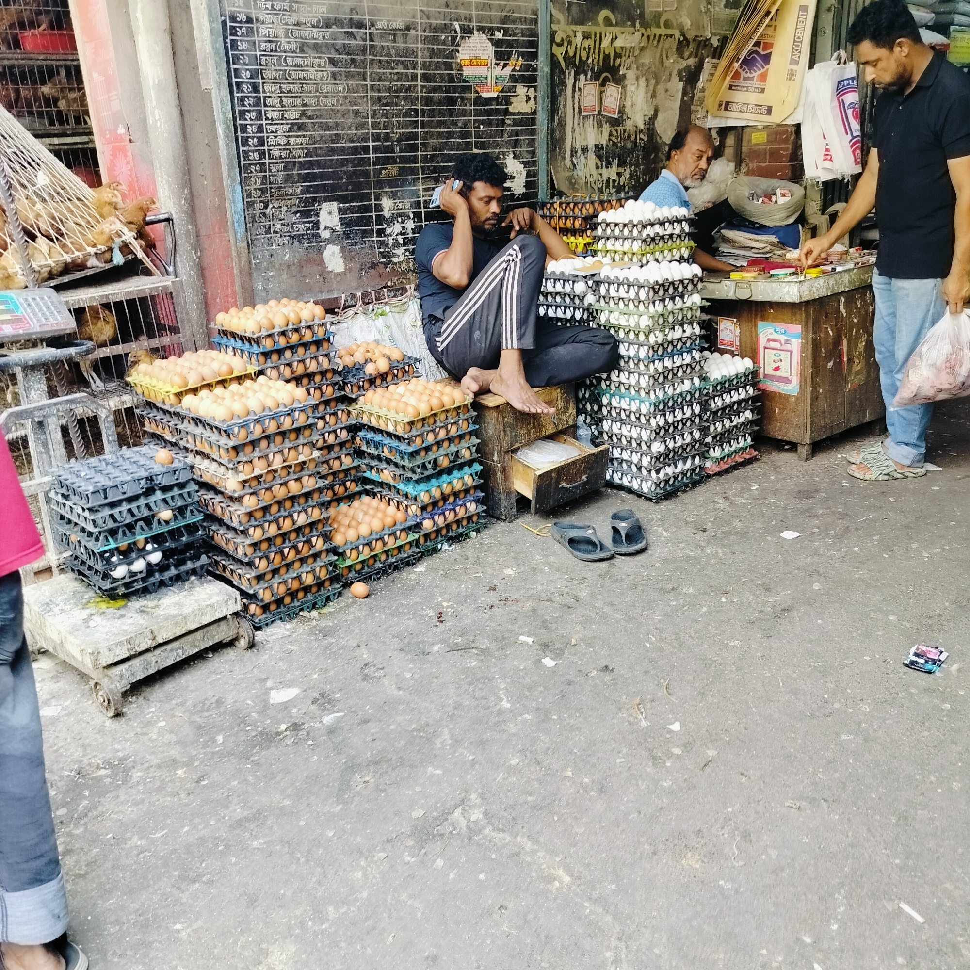 This image depicts a street scene in Dhaka, Bangladesh, likely a marketplace. The scene features multiple individuals and focuses on the sale of eggs. In the foreground, stacks of egg trays filled with brown and white eggs dominate the left and center. One man, barefoot, sits on a small wooden box amidst the eggs. He is dark-skinned, wearing a black t-shirt with striped black pants, and appears to be on a cell phone call. Behind him is a dark board covered in Bangla script, likely a price list. Another man in a light blue shirt stands behind a counter, and a third man in a black t-shirt and jeans stands to the right, holding a plastic bag. A cage containing chickens is visible in the upper left. The foreground is dominated by a concrete pavement. Trash and debris are scattered on the ground, including a discarded pack of playing cards. Bags of cement can be seen.