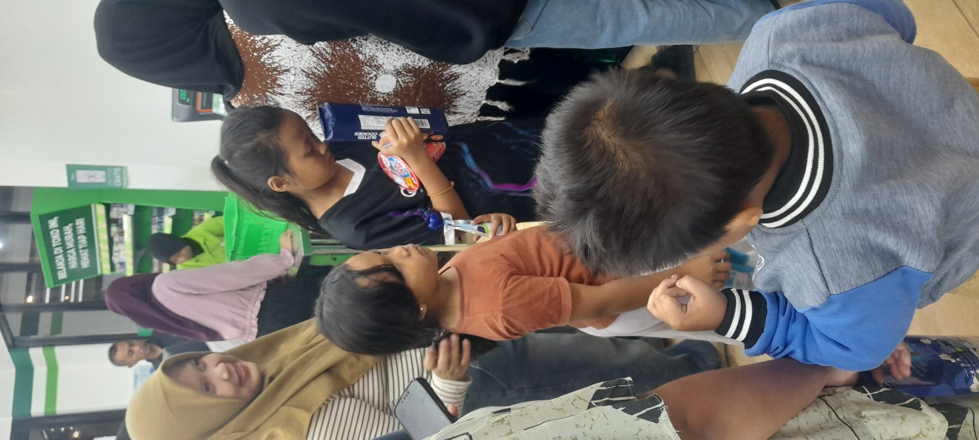 In Kragilan, Indonesia, a group of people are gathered in what appears to be a retail store, possibly a minimarket, with green shelves in the background displaying products. A woman wearing a tan hijab and a striped shirt smiles at the camera, holding a dark smartphone. Next to her, a young girl in a black t-shirt with a colorful graphic is holding a blue box of cookies and a red cylindrical object. Another young girl, facing to the right, wears an orange t-shirt and white pants and holds a small white and blue object. In the foreground, the back of a young boy's head is visible; he is wearing a grey and blue sweatshirt with black and white striped trim. The overall scene suggests a casual shopping or waiting situation within a commercial setting.