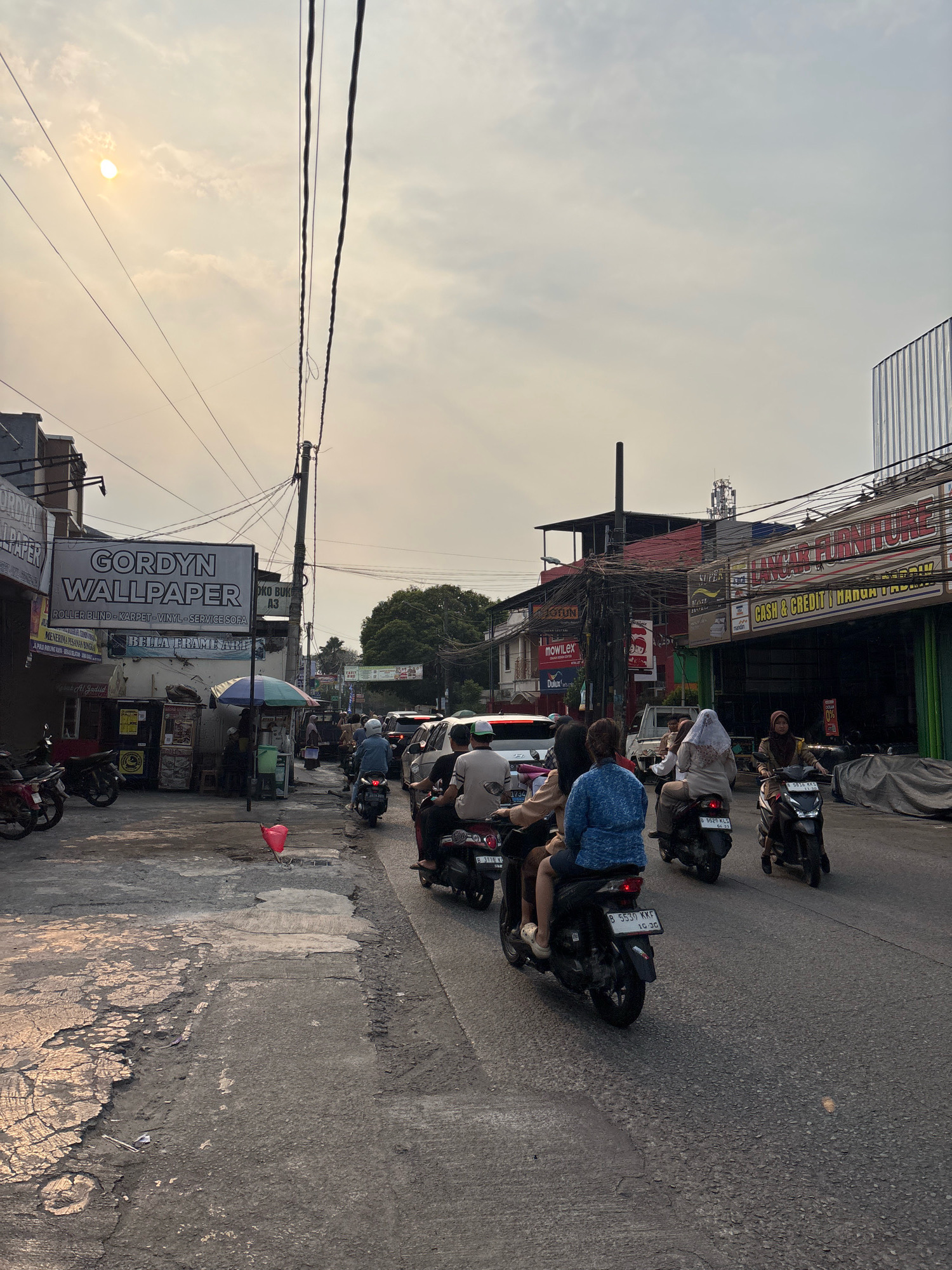 The image captures a busy street scene in Bekasi, Indonesia, likely during late afternoon, as indicated by the low sun casting a warm glow and long shadows. The foreground features a cracked and weathered asphalt road, with some areas showing exposed concrete. To the left, a sidewalk area is occupied by parked motorcycles and street-side stalls, one displaying a prominent sign for "GORDYN WALLPAPER" along with "ROLLER BLIND - KARPET - VINYL - SERVICE SOFA" and "BELLA FRAME ART". Further down the sidewalk, a small red plastic bag lies on the ground.
The street is filled with traffic, predominantly motorcycles, with several riders and passengers visible. Some individuals are wearing helmets, while others are not. Several cars are also present in the traffic jam. Buildings line the right side of the street, displaying various storefronts with prominent signage, including "HONGKART FURNITURE" and "CASH & CREDIT HONGKART". Overhead, a dense network of electrical wires crisscrosses the sky, supported by utility poles. In the distance, trees and more buildings are visible, along with a telecommunications tower. The sky is overcast with some diffused sunlight. The overall impression is one of a bustling urban environment in Bekasi, Indonesia.