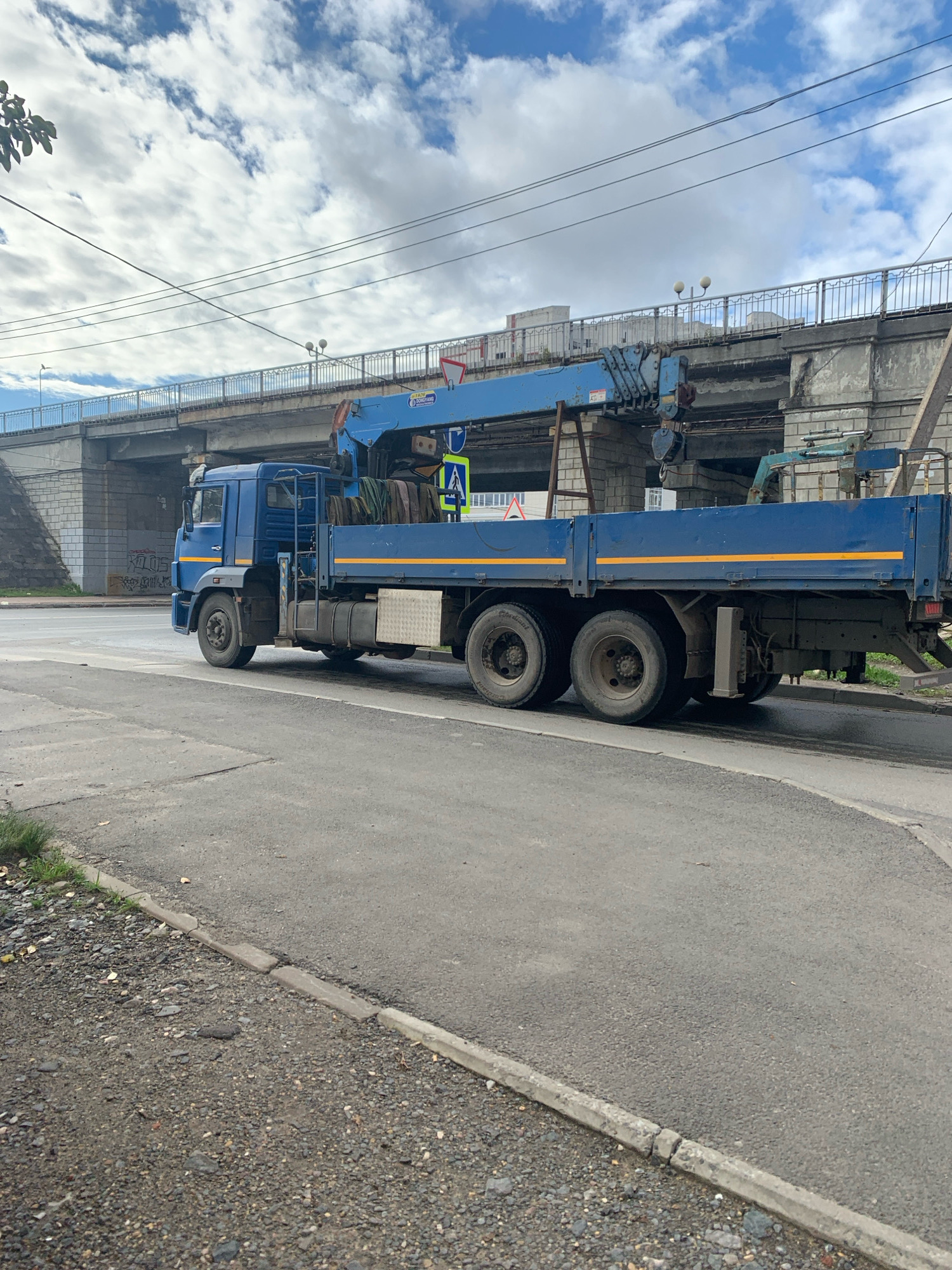 A blue flatbed truck with a crane is parked on a paved road next to a gravel shoulder. The truck has a long flatbed with yellow reflective tape along the sides and a blue crane extended over the bed, holding several bundles of what appear to be cables or pipes. The truck has six wheels, with a double axle at the rear. In the background, a concrete overpass or bridge with a metal railing and streetlights spans across the scene. The sky above is cloudy with patches of blue. The area appears to be an urban environment, possibly on the outskirts of Arkhangelsk, Russia, given the type of infrastructure and vehicle. The road has visible lane markings, and there is some graffiti on a concrete wall to the left of the truck. There are no people visible in the image.