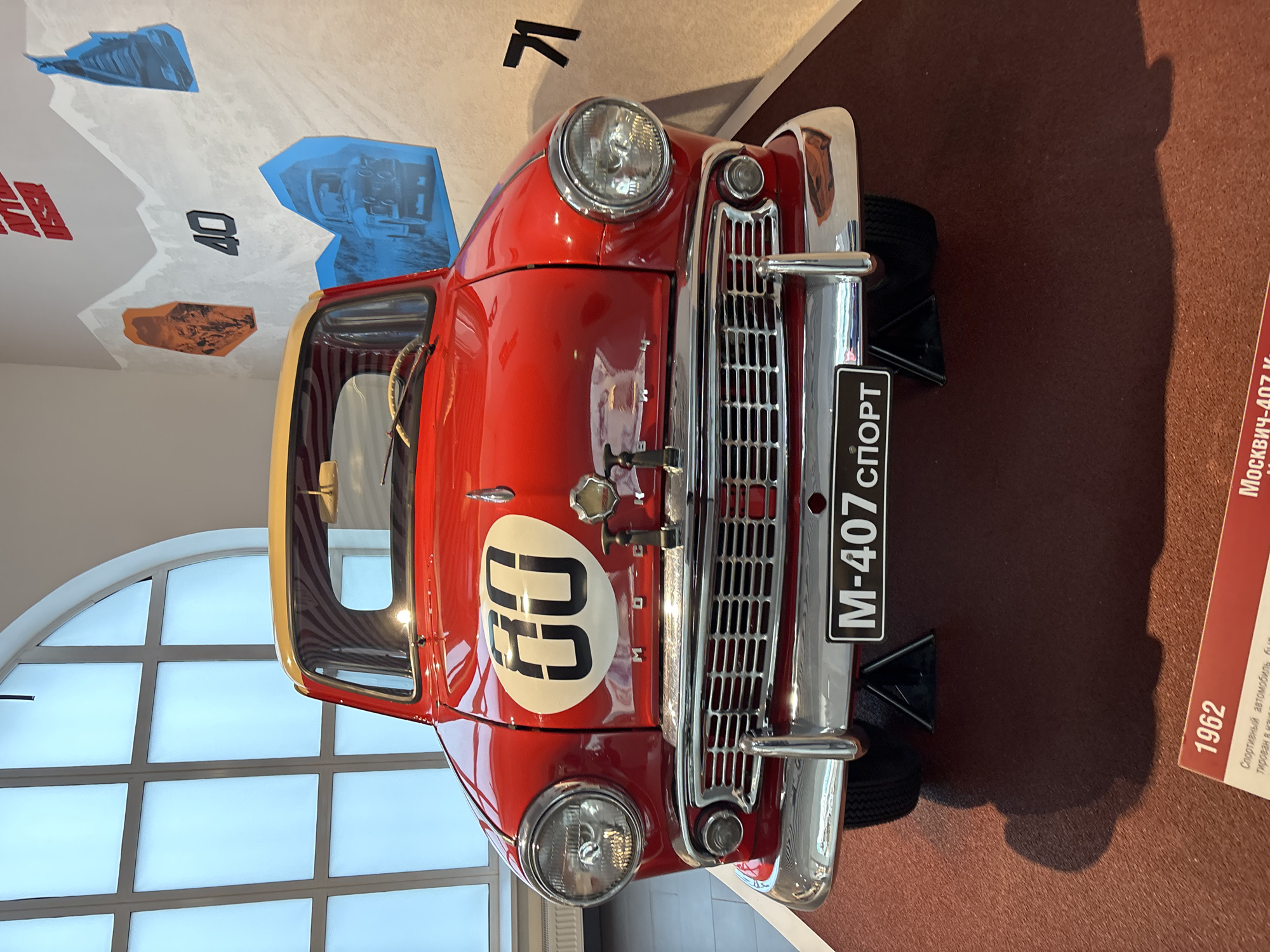 This is a head-on, eye-level shot of a red and yellow Moskvich-407 Sport car, dated 1962, on display at the Military Automotive Museum in Verkhnyaya Pyshma, Russia. The car features a prominent white circle on its hood with the number "60" in black. A black license plate with "M-407 СПОРТ" is mounted on the chrome bumper. The car is positioned on a red carpeted platform, with black triangular supports visible beneath the front wheels. Behind the car, a wall is decorated with graphic art, including mountain silhouettes, a large number "40", and images of older vehicles. To the left of the car, a large, arched window with frosted panes is visible. A portion of a red informational plaque is visible in the lower left corner of the image, displaying the year "1962" and some Cyrillic text. The overall environment appears to be an indoor museum exhibit.