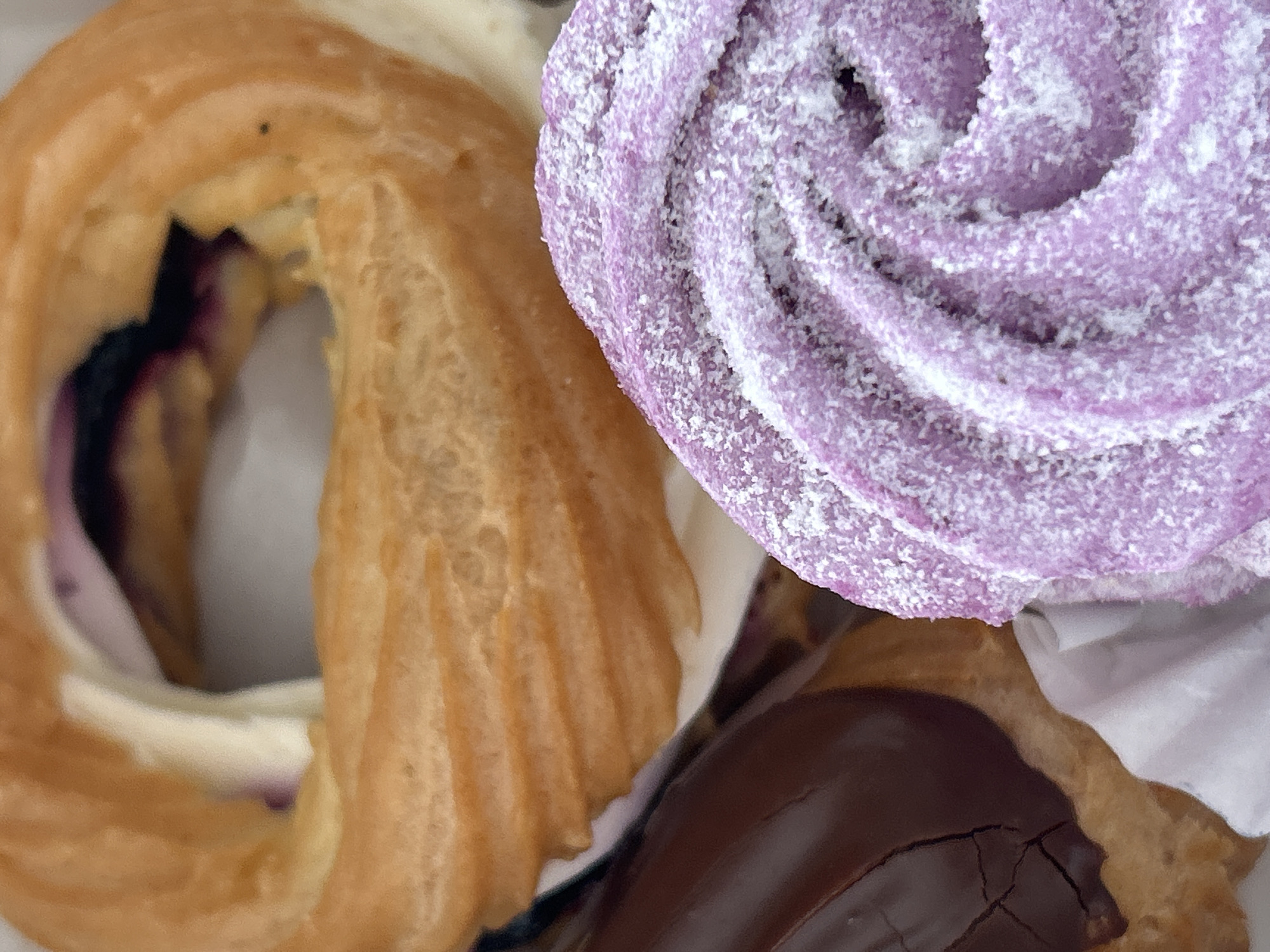 The image shows a close-up, overhead view of three pastries on a white surface. The pastries are arranged in a cluster. On the left is a ring-shaped pastry with a ridged, golden-brown exterior and white cream filling peeking out. Within the opening of this pastry, a dark purple filling, likely fruit, is visible. To the right of this is a swirl-shaped pastry, a vibrant purple, covered in a fine white powder, possibly powdered sugar. Below these two pastries, a portion of a dark chocolate-covered pastry is visible, with a glossy finish and some cracks in the chocolate. The lighting suggests it might be indoor lighting. There is no visible text in the image. The context provided suggests the location is Марковский, Russia, but there are no specific visual cues in the image to confirm this.