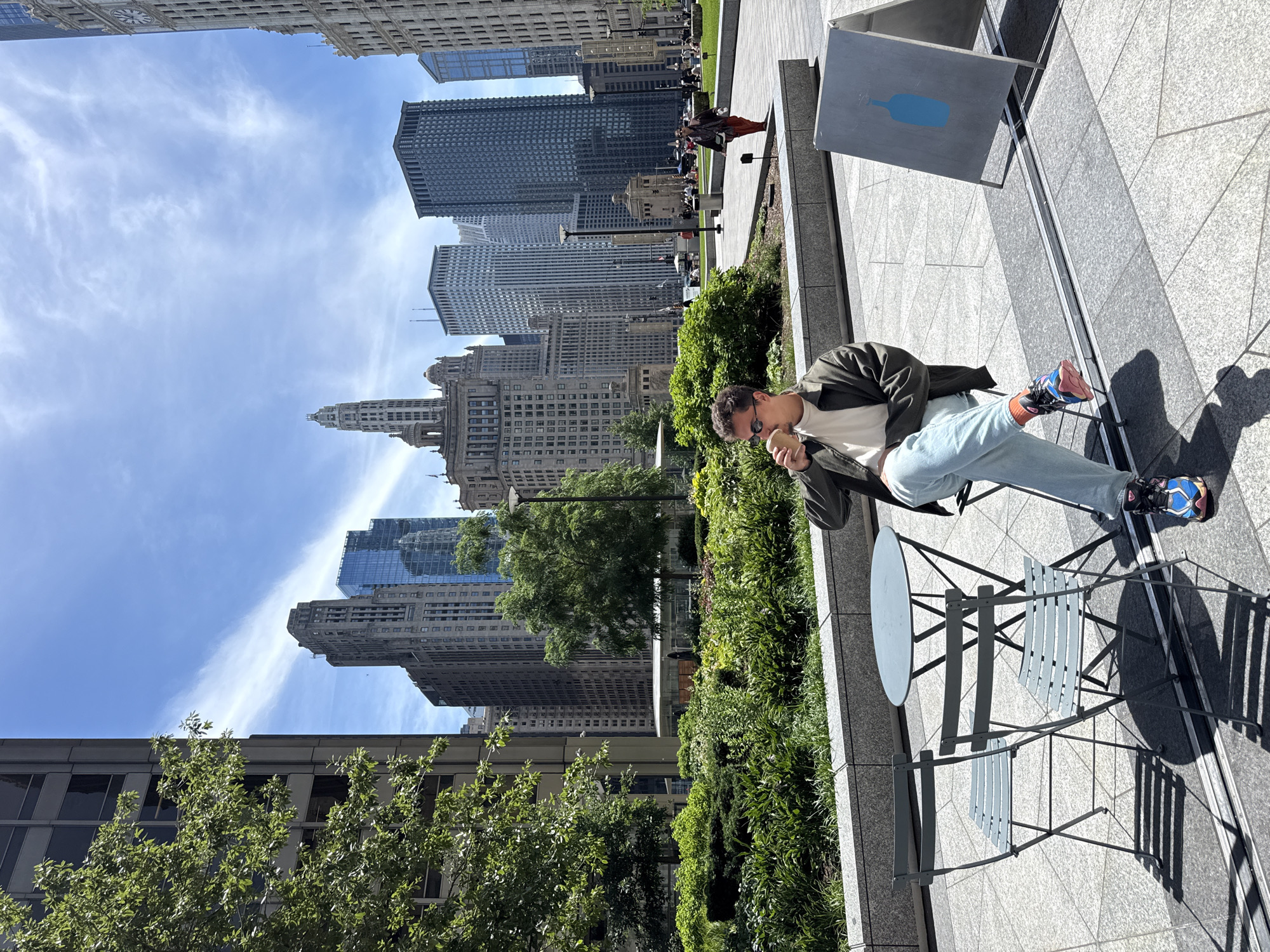 A man is seated outdoors at a table and chairs, holding a cup to his mouth. He is wearing a dark green jacket over a cream-colored shirt, light blue pants, and colorful sneakers. The scene is set in a plaza with a backdrop of tall buildings, indicative of a city environment. To the right of the man is a blue bottle graphic on an A-frame sign. In the background, there is a lush green park area with trees and manicured bushes, and other people are visible in the distance walking and sitting. The sky is bright blue with scattered white clouds, suggesting a sunny day. The architecture of the surrounding buildings is a mix of older, ornate structures and modern glass skyscrapers. This appears to be a public space in Chicago, likely within the Loop or a similar downtown area, given the density of the architecture and the presence of urban green spaces.