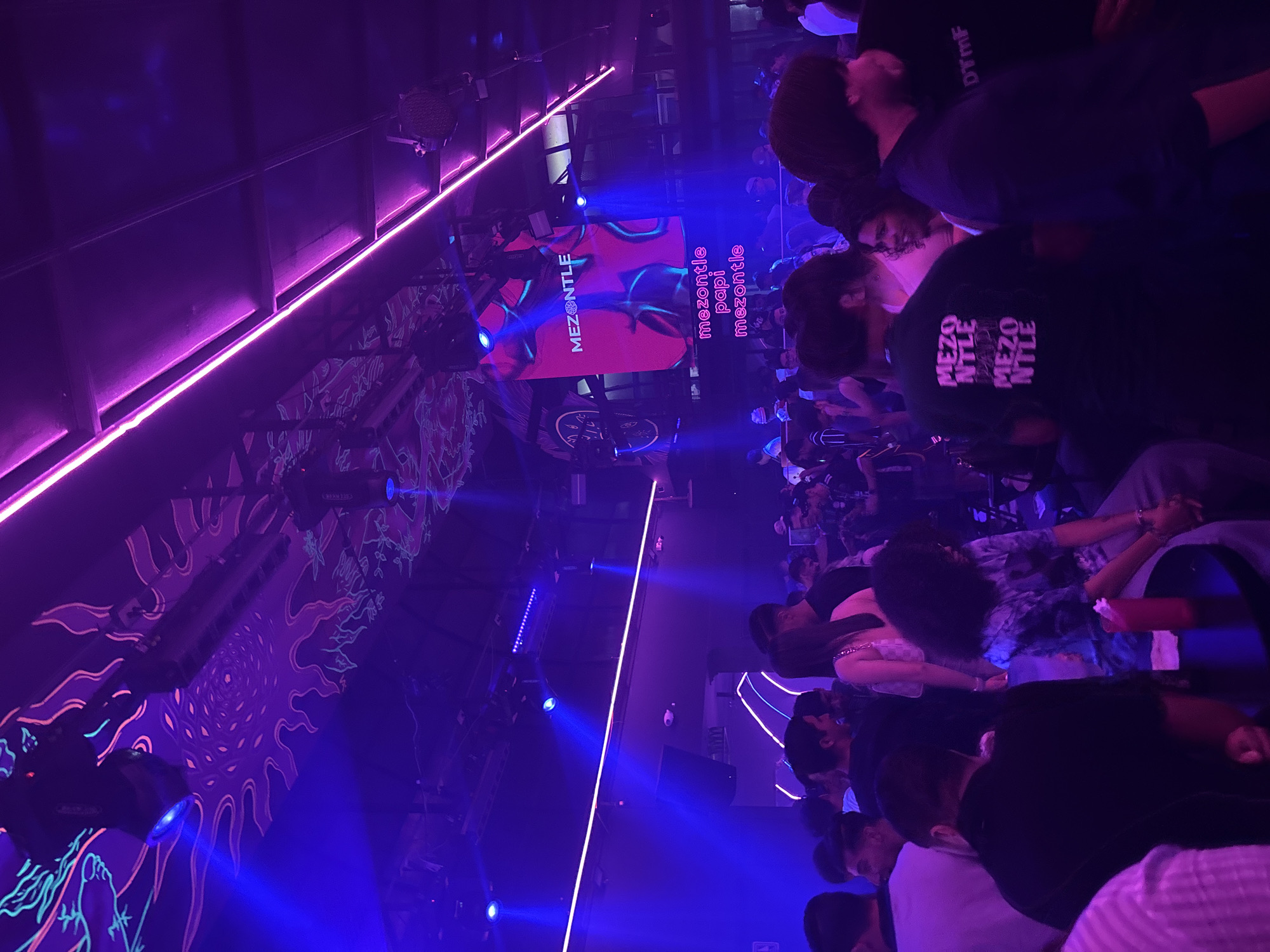 The image shows an indoor event, possibly a nightclub or concert in Veracruz, Mexico. The scene is illuminated with bright neon blue and purple lights. A large screen displays the word "MEZONTLE." Many people are visible in the audience. Some are wearing dark clothing, and one has a shirt with "MEZONTLE" printed on it. The crowd seems relaxed, possibly enjoying the event. The room's decor features psychedelic patterns. Overhead, lighting fixtures and stage equipment are visible. The photograph is taken from a slightly elevated and tilted perspective.
