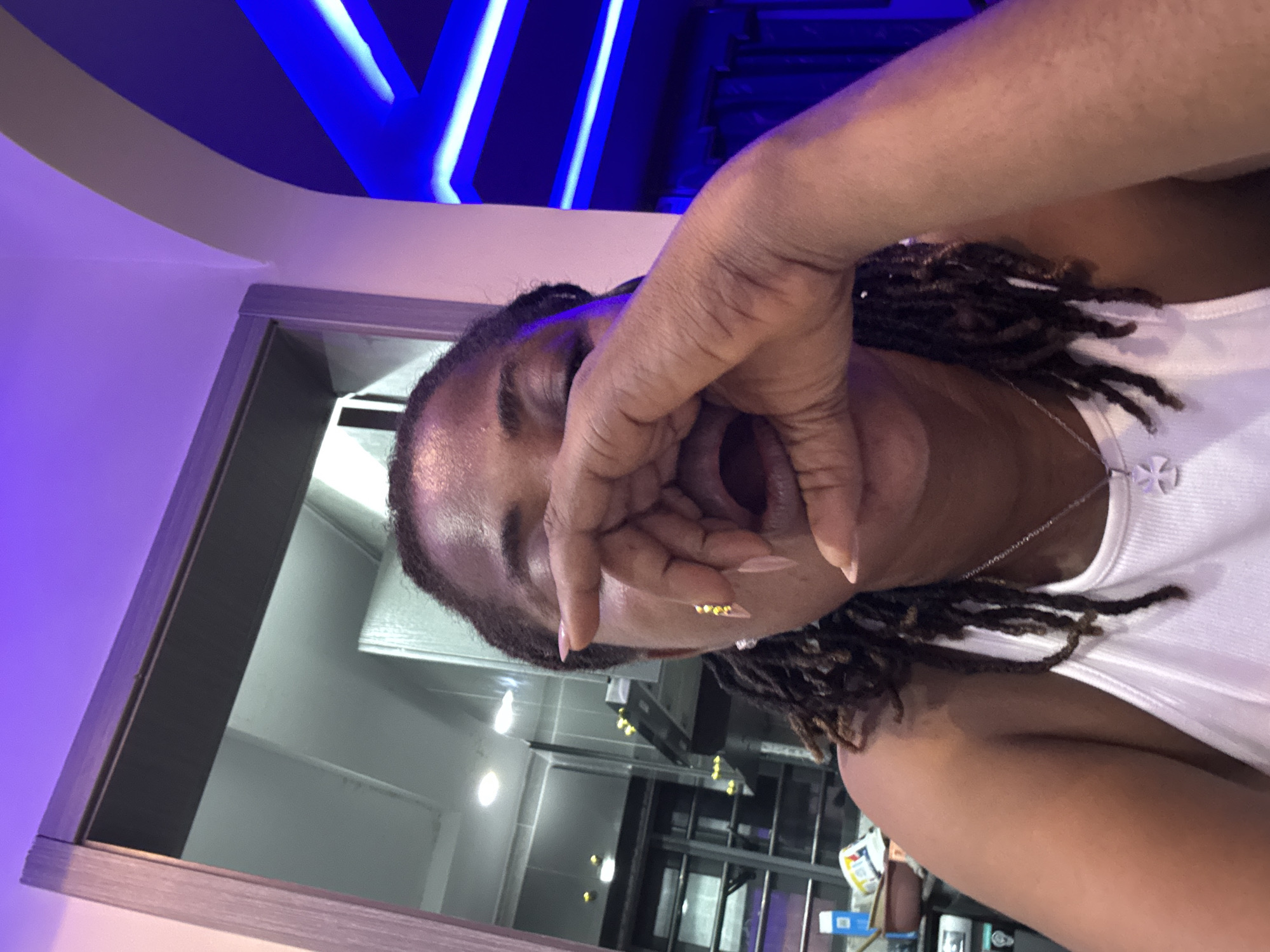 The image shows a person with dark skin, wearing a white tank top and a silver necklace with a cross pendant. Their face is partially obscured by their hand, which is covering their mouth and nose. The person has dreadlocks and is wearing a pearl earring and what appear to be gold embellishments on their fingernails. The background suggests an indoor setting with purple and blue lighting. To the left, a doorway leads to a room with what looks like kitchen appliances and shelves. To the right, the ceiling features illuminated blue stripes. The scene is likely in Ajara, Nigeria.