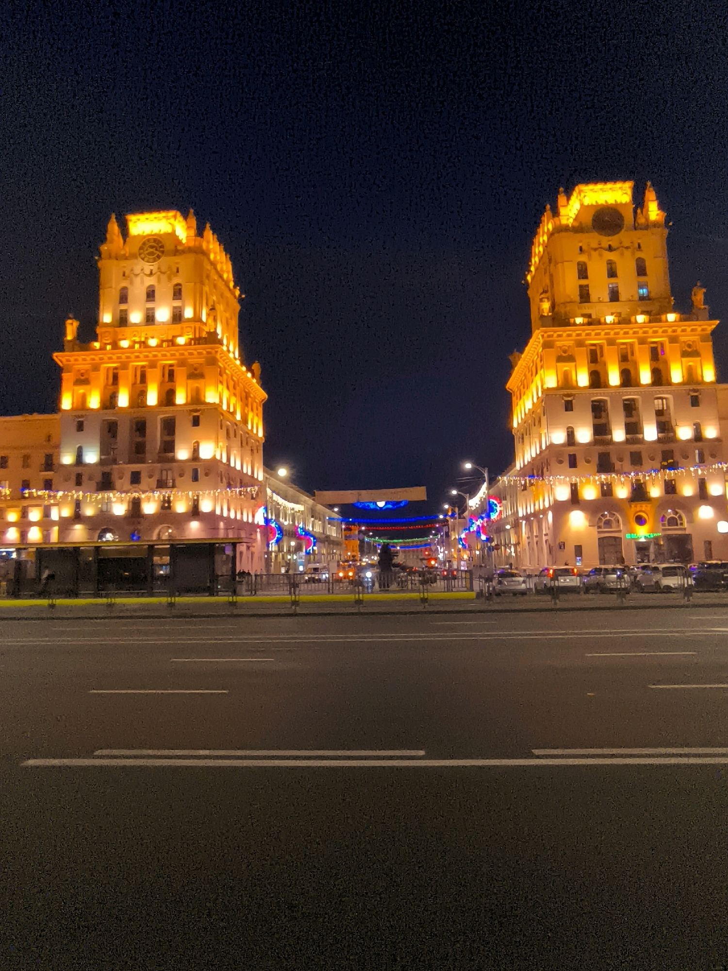 The image shows the Gates of Minsk in Minsk, Belarus, at night. The iconic twin buildings are illuminated with bright orange lights, highlighting their architectural details and the large clocks on their facades. The sky above is a deep dark blue, indicating the late hour. In front of the buildings, a wide, multi-lane road is visible, with white lane markings. Several cars are parked or driving on the road. Decorative lights are strung across the street between the buildings, adding a festive atmosphere. A bus stop shelter is present on the left side of the image, and some streetlights are visible. The overall scene is a well-lit urban landscape at night.