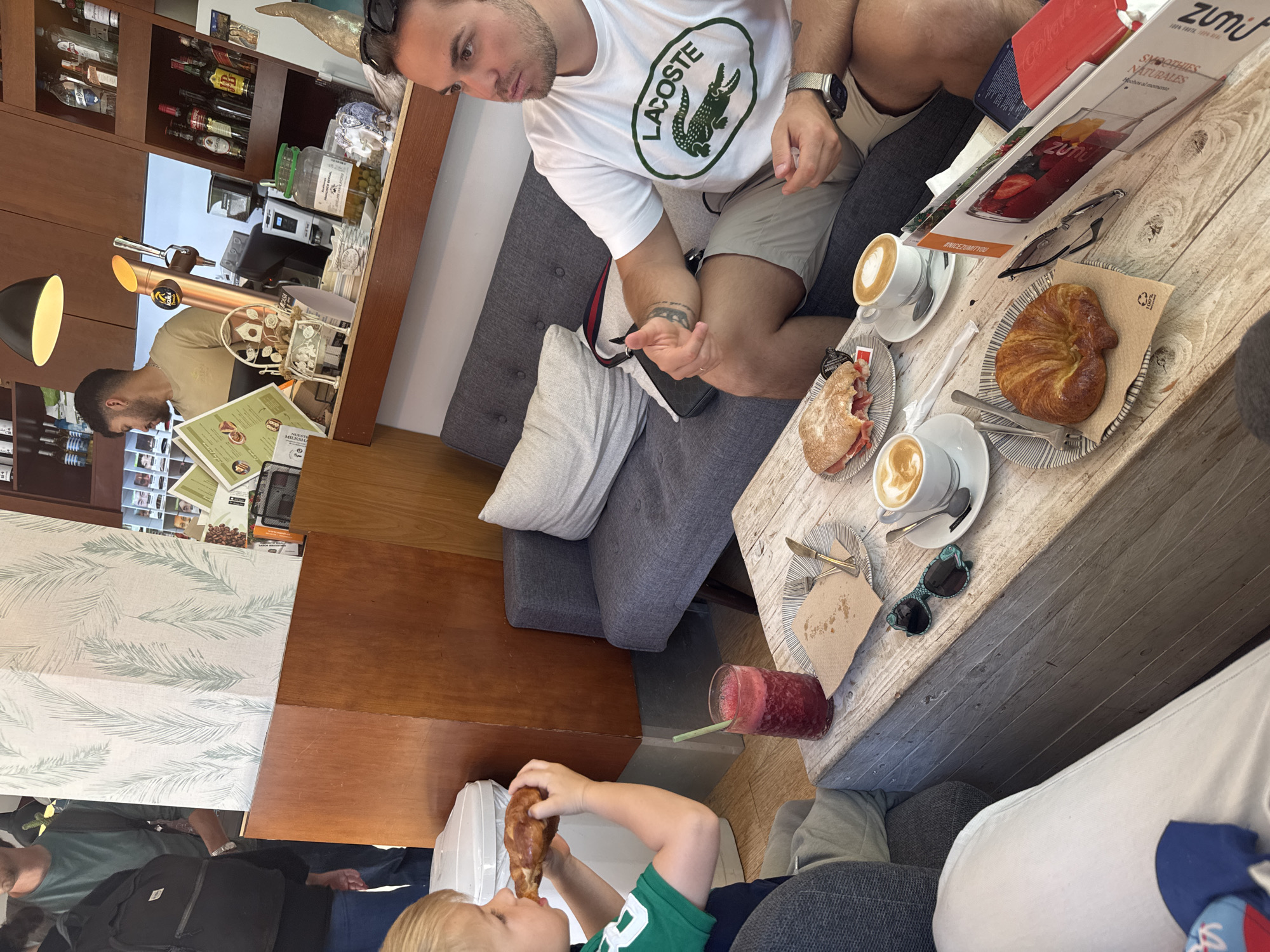 The image captures a scene in a café in Valencia, Spain. A man in a white Lacoste t-shirt and shorts sits on a grey cushioned bench. He is looking at something in his hand, possibly a mobile phone. On the table in front of him, there are two cups of coffee, a sandwich, a croissant, a smoothie, sunglasses, and a menu. A child wearing a green t-shirt is sitting next to the table, eating a croissant. A waiter is visible in the background, serving behind a counter with a display of food and beverages. Shelves with bottles of alcohol are above the counter.
