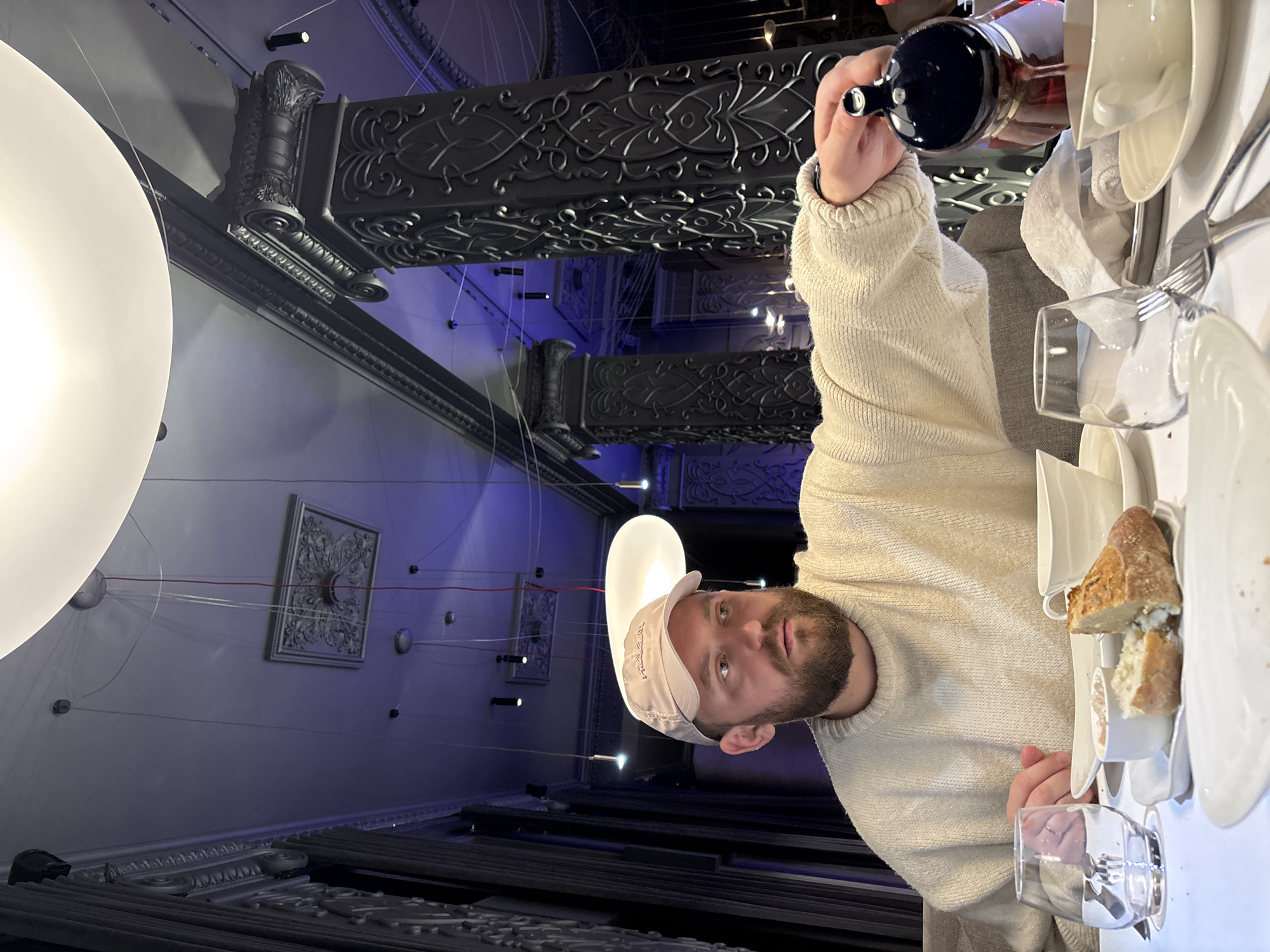 The image captures a male individual seated at a dining table in an upscale restaurant, identified as Malina Bar & Restaurant, located at ul. Lena, 118, Krasnoyarsk, Russia. The man, a bearded Caucasian, wears a beige baseball cap and a cream-colored knitted sweater. He is actively pouring a dark liquid from a black, globular teapot into a white ceramic teacup positioned on a matching saucer.
The dining table is covered with a white tablecloth and set with white ceramic dinnerware, including a main plate, a smaller plate holding a piece of baguette bread and a small bowl with a white spread, a stemless clear glass, and silverware. In the background, the restaurant's interior is characterized by dark, ornate columns featuring intricate carvings and classical capitals with volutes and acanthus leaves. The light-colored ceiling displays decorative molded panels and a central rosette, from which multiple suspension lights hang, including large, flat, oval white luminaires and smaller cylindrical spotlights. Blue accent lighting is visible along the ceiling and walls, contributing to the dramatic ambiance. Dark, textured wall coverings or drapes are visible on the far left. No other persons are clearly visible, but the table setting implies multiple diners.
