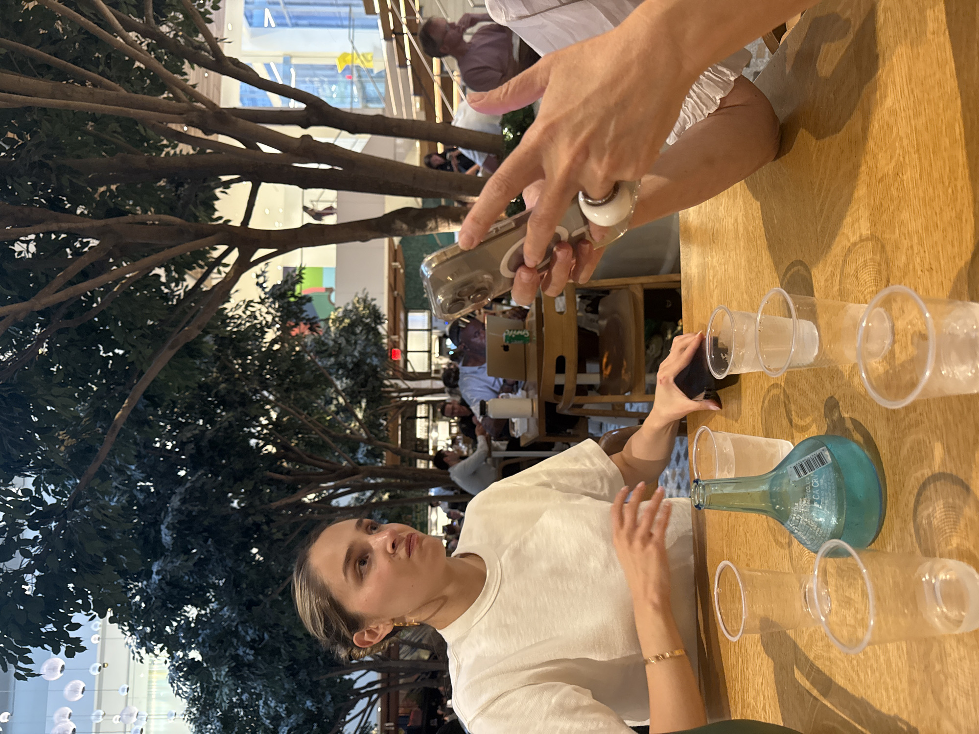 The image captures an indoor scene with a focus on a table laden with plastic cups and a turquoise glass bottle containing a blue liquid. In the foreground, a woman with her hair tied back is looking to her left, her expression suggesting deep thought or perhaps mild annoyance. She is wearing a white t-shirt and a delicate bracelet. Her hands are resting on the table, with one hand blurred as she gestures. To her right, another person's hands are visible, holding a smartphone in a clear case and wearing a ring on their index finger. This person is also wearing a white top with ruffled details. The table is made of light-colored wood, and the surrounding environment appears to be a restaurant or cafe with trees and architectural elements visible in the background. The overall impression is a casual gathering at a table, possibly in a bustling urban setting.