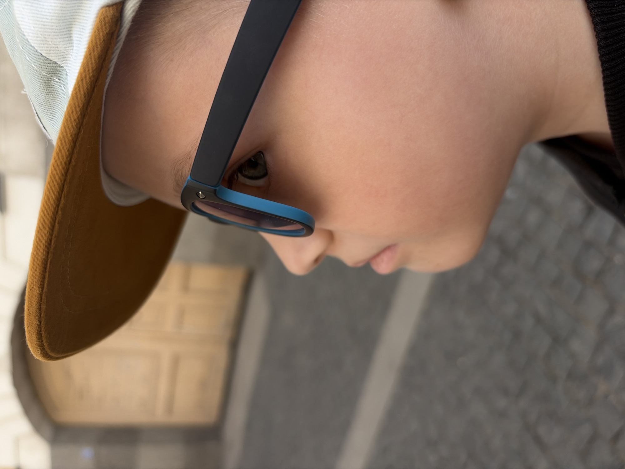 Here's a forensic description of the image:
**Subject:** A close-up, slightly angled view of a child's face.
**People:** A child wearing glasses and a baseball cap.
**Objects:** Glasses with black frames and blue rims, a baseball cap with a mustard-colored bill and light-colored crown, a blurred doorway, and blurred street pavement.
**Environment:** The background suggests an outdoor location, likely a street or sidewalk, and a building doorway that might be in the city of Saint Petersburg, Russia.