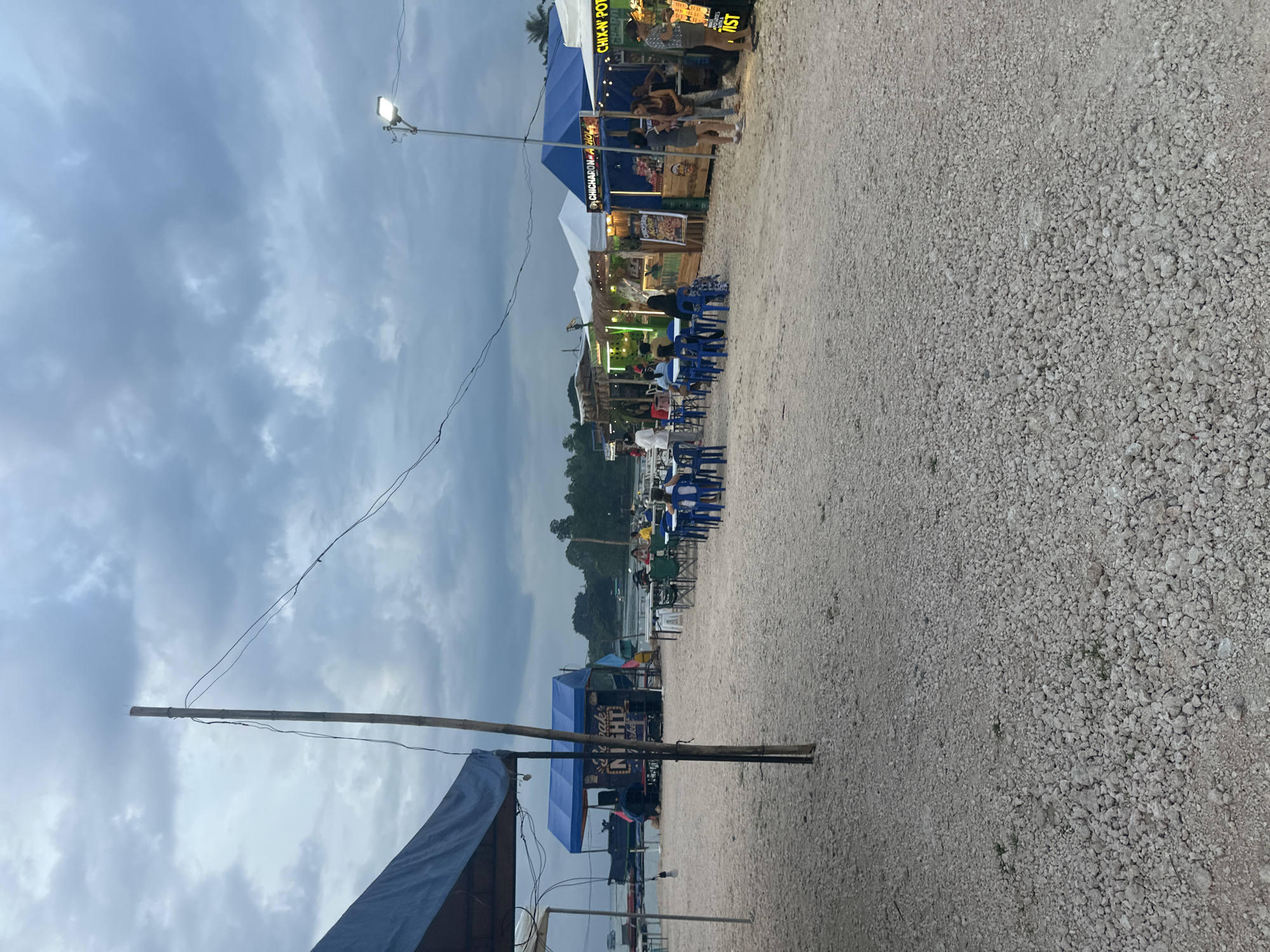 An outdoor market with food stalls and seating areas is set up on a gravelly beach in Samal, Philippines, under a cloudy sky. Several stalls are visible, some with blue awnings and signage, including one with the words "CHIX-N POTA" and another reading "CHICHARON HOJA." People are seen standing and sitting at tables, some eating or conversing. A street light illuminates the scene. The background shows a body of water with boats and lush greenery. A wooden pole with wires runs through the foreground. The overall atmosphere suggests an evening gathering or dining experience.