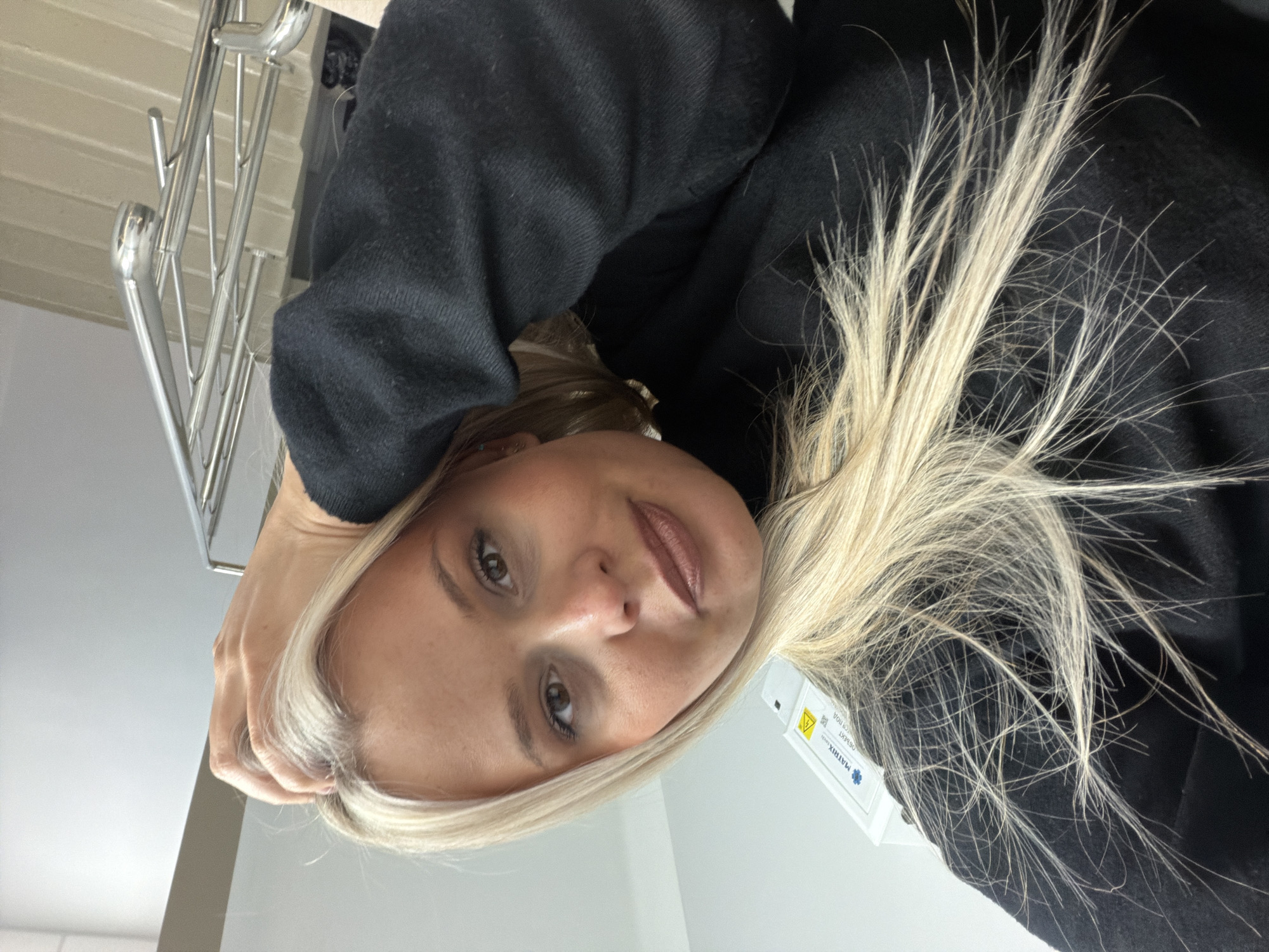 A woman with blonde hair is looking at the camera. She is wearing a black sweater and a medical device is attached to her hair. The background is a white wall and a metal railing. The location is not specified in the image and cannot be determined without additional information.
