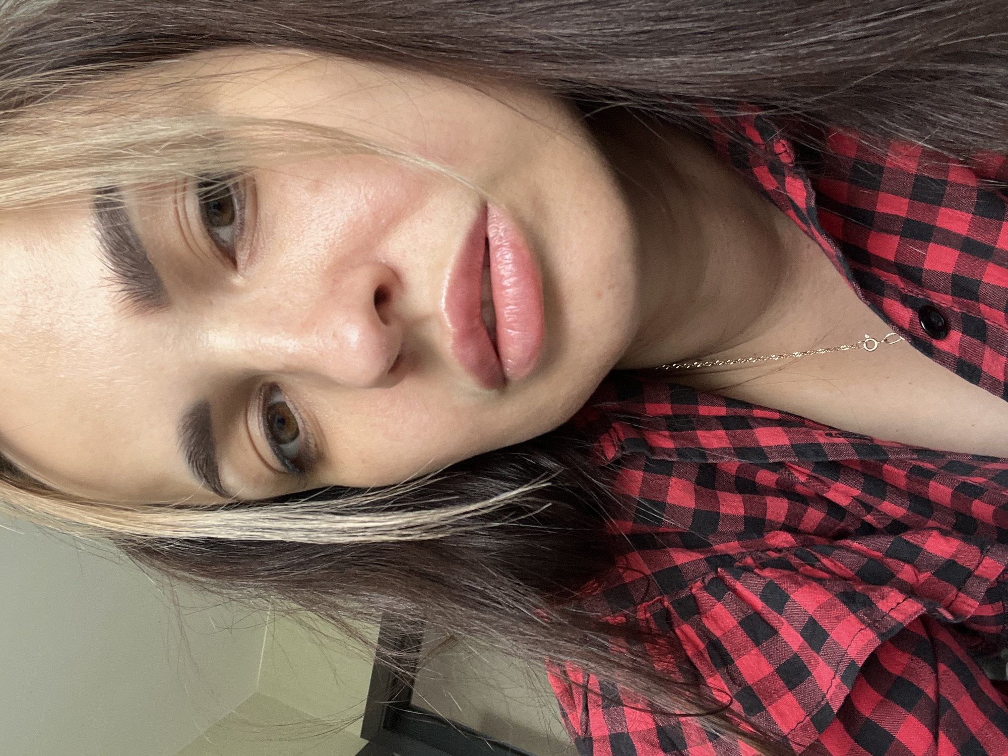 A close-up, rotated portrait features a woman's head and upper torso, viewed from a slightly upward angle. The image is oriented sideways, with her head tilted towards the left side of the frame when viewed upright. She has long, dark brown hair, with distinct light blonde highlights or streaks framing her face on the left side. Her right eye is partially visible, showing a brown iris, and her eyebrows are well-defined. Her lips are full, glossy, and a natural light pink or nude shade, slightly parted.
She is wearing a casual red and black plaid (gingham) button-up shirt. A thin, gold-colored chain necklace with a small clasp is visible around her neck. The background appears to be an indoor setting, with a plain, light-colored wall visible in the upper left corner and a dark, rectangular object, possibly part of furniture or a door frame, in the lower right. The lighting is soft and even, creating a clear and well-lit image of her face. Her expression is calm and neutral. There is no visible text in the image.