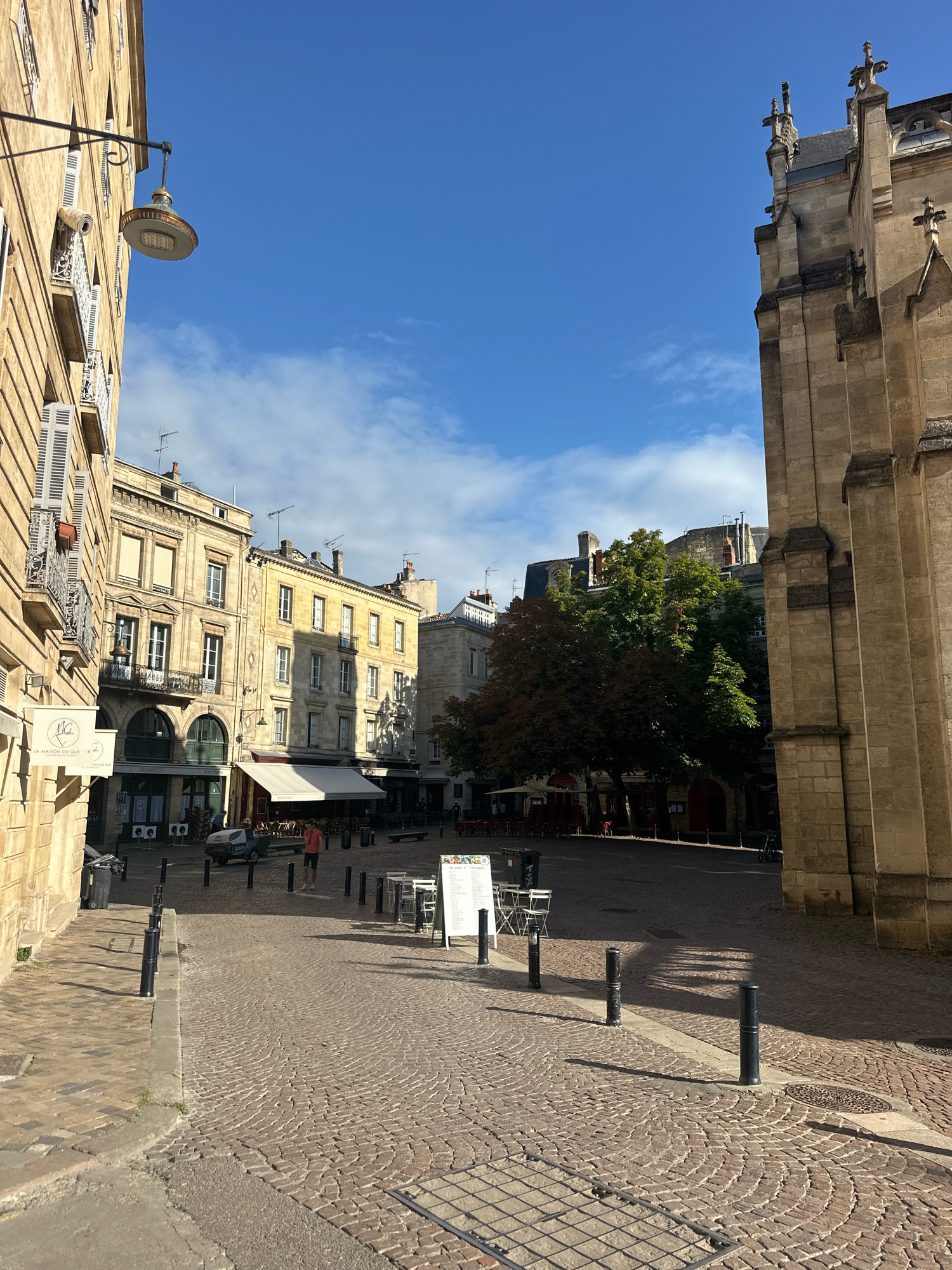 The image depicts a street scene in Bordeaux, France, likely within the historic center, given the architectural style. In the foreground, a cobblestone street is visible, with a paved sidewalk to the left. The street is lined with traditional European buildings characterized by their stone facades, arched windows, and shutters. A prominent, ornate street lamp extends from the building on the left.
Further into the scene, there is an open plaza area with several tables and chairs set up, suggesting an outdoor seating area for cafes or restaurants. Some of these tables have umbrellas. A white menu board stands in the plaza. A few people are present, including one individual in the middle ground wearing a red t-shirt and shorts, walking away from the camera. A small utility vehicle is parked in the mid-ground.
To the right of the frame, a large, imposing stone building with Gothic architectural elements, possibly a church or cathedral, dominates the view. The sky is blue with scattered white clouds. Trees with green and reddish foliage are visible in the mid-ground, casting shadows on the plaza. The overall impression is of a sunny day in a European city, with a blend of historical architecture and urban amenities.