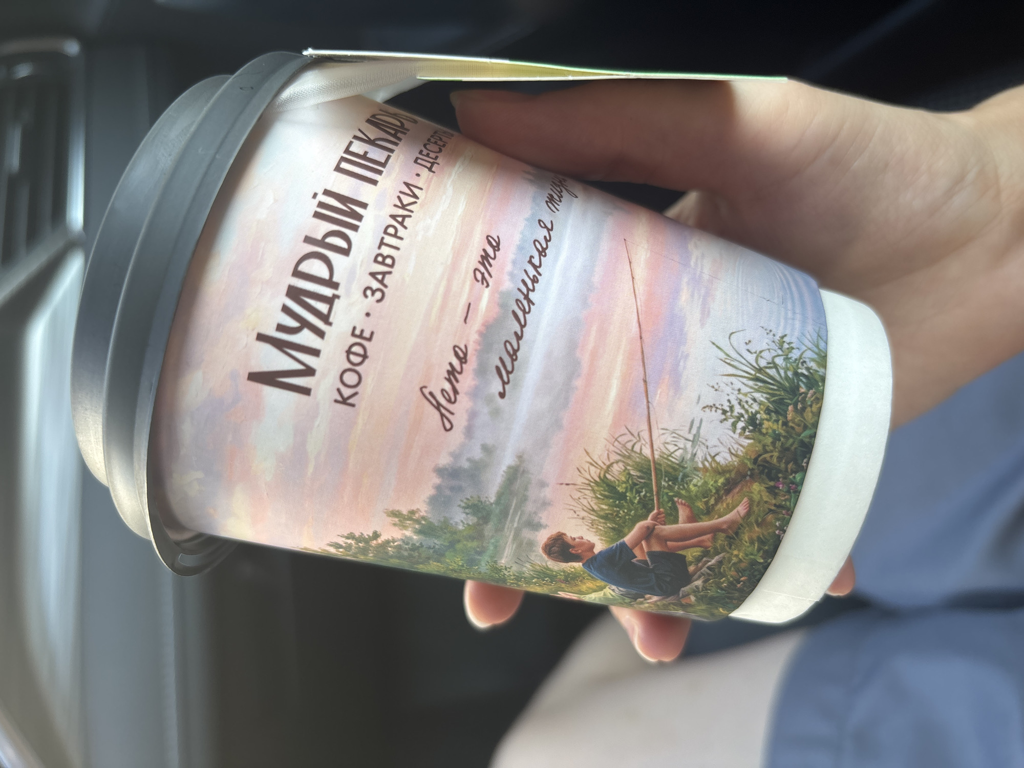 A person's hand holds a paper coffee cup with a lid. The cup is decorated with a painting of a young boy fishing by a misty lake at sunrise. The text on the cup reads "МУДРЫЙ ПЕКАРЬ" (Wise Baker) and "КОФЕ • ЗАВТРАКИ • ДЕСЕРТЫ" (Coffee • Breakfasts • Desserts). Below the painting, handwritten text says "Лето - это маленькие тихие" (Summer is the little quiet). The background is blurred, showing an interior environment, possibly a car. There is no visible indication of the city of Naberezhnye Chelny, Russia.