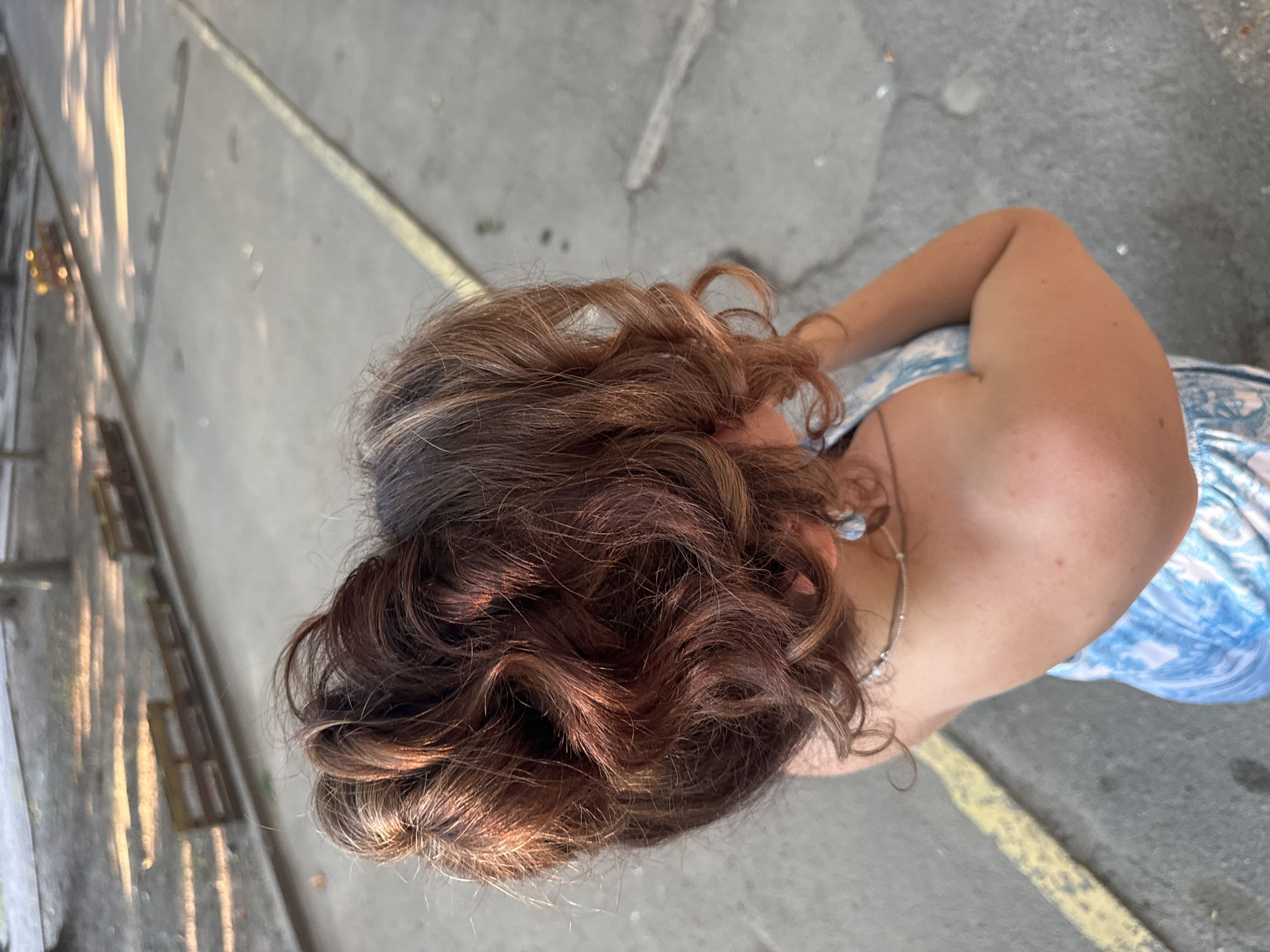 This is a top-down, slightly angled outdoor photograph of a person from the chest up. The subject has curly, brown hair that is lit by sunlight, highlighting copper and auburn tones. They are wearing a light blue and white patterned dress and a silver necklace with a small pendant. A portion of their right arm and shoulder is visible, and they are wearing a circular earring in their visible left ear. The background is a gray asphalt surface with a yellow painted line. Sunlight casts shadows on the ground from an unseen object on the left side of the frame. The image appears to have been taken during the daytime, with bright sunlight suggesting fair weather. The scene is likely a public outdoor space, possibly a street or walkway, given the asphalt and painted line. There is no visible text in the image.