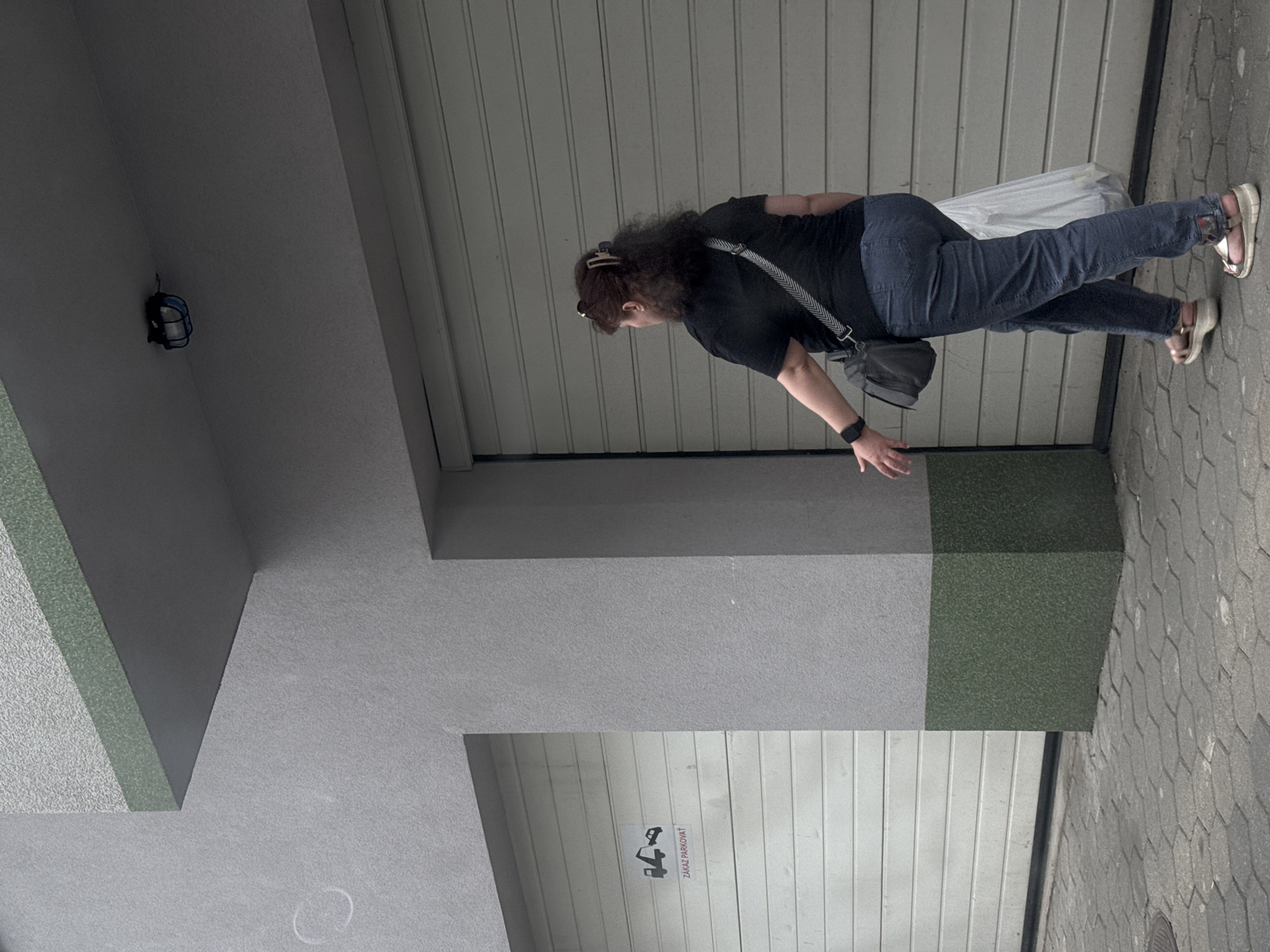A woman with curly brown hair, wearing a black t-shirt, blue jeans, and sandals, walks away from the camera. She is carrying a grey shoulder bag and a white plastic bag. She is walking on a paved walkway in front of a building with grey walls and a green-bottomed pillar. To her left are two large garage doors, one of which has a "No Parking" sign. The scene appears to be a street in Bratislava, Slovakia. The lighting suggests it is daytime, but overcast.