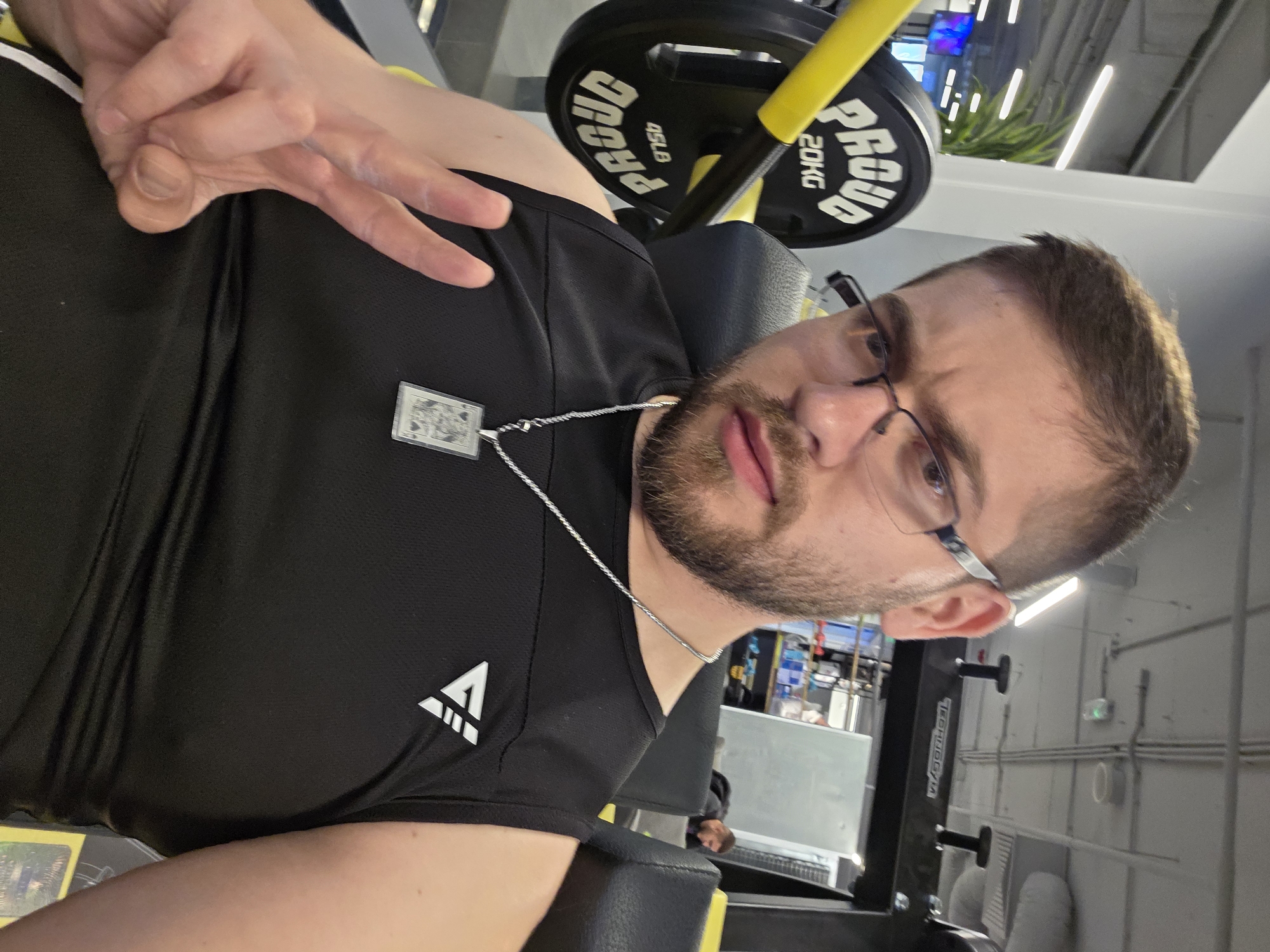 This is a slightly tilted, medium close-up shot of a man in an indoor gym environment, located in Plovdiv, Bulgaria. The man, with short brown hair, a neatly trimmed beard, and dark-rimmed glasses, looks directly at the camera with a neutral expression. He is wearing a black short-sleeved t-shirt featuring a white triangular logo on the left chest. Around his neck, he wears a silver chain with a rectangular pendant that resembles a playing card, possibly the King of Spades. His left hand is raised, forming a peace or victory sign with his index and middle fingers.
The man appears to be seated or resting on a piece of black and grey gym equipment, possibly a bench or machine seat. In the upper right background, a black weight plate is prominent, with yellow lettering that reads "PROUD," "45LB," and "20KG," attached to a yellow bar. Further in the background, typical gym elements are visible, including fluorescent strip lighting on the ceiling, some artificial greenery, and other workout machines. One black machine in the distance bears the partial text "TECHNOGYM." The lighting suggests an indoor setting, and the man seems to be posing for a casual photo or selfie during a workout break.