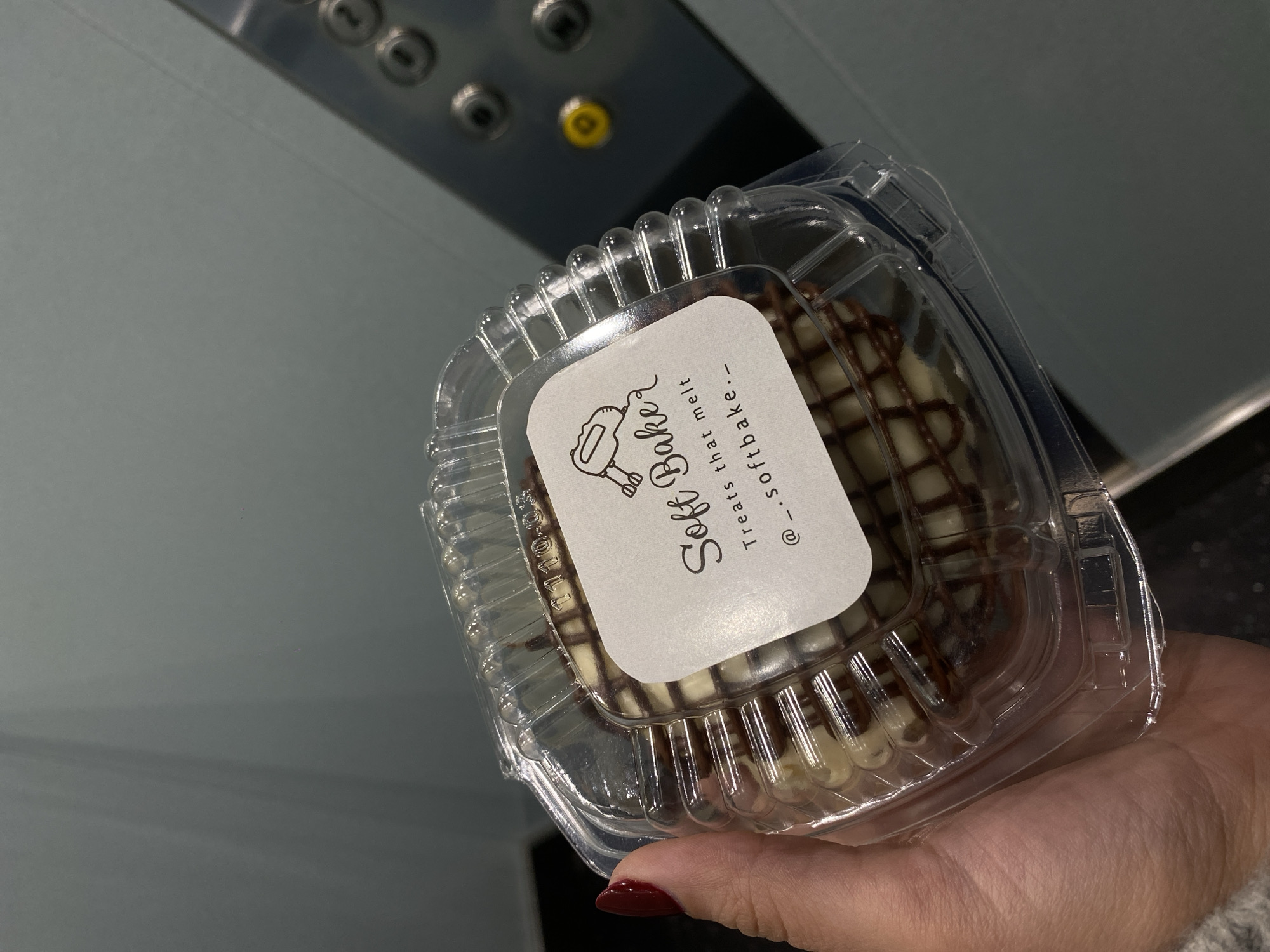 A person with a red nail is holding a clear plastic clamshell container with a baked good from "Soft Baker - Treats that melt - @_.softbake._". The background is a metallic elevator wall with buttons. The image was likely taken in Puebla, Mexico, given the baker's Instagram handle.