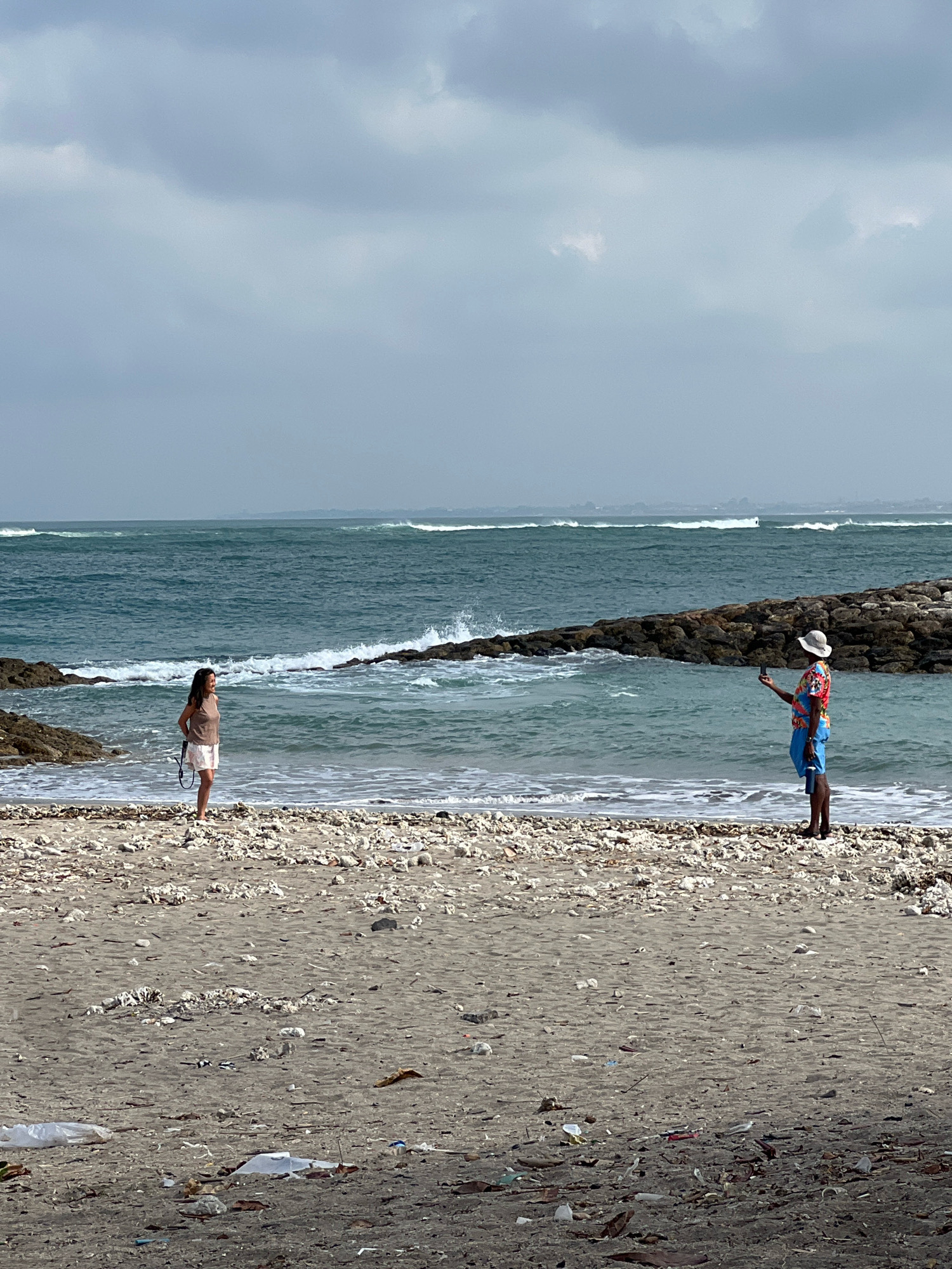 An overcast day at a Kuta, Indonesia, beach. Two individuals are on the sandy shore, with the ocean and waves in the background. A woman in a brown tank top and white skirt stands to the left, facing the sea. To the right, a man in a colorful shirt and blue shorts holds up a phone, seemingly taking a photo or video. The beach itself is covered with debris, including coral, driftwood, and plastic waste. A rock jetty stretches into the ocean on the right side of the frame.