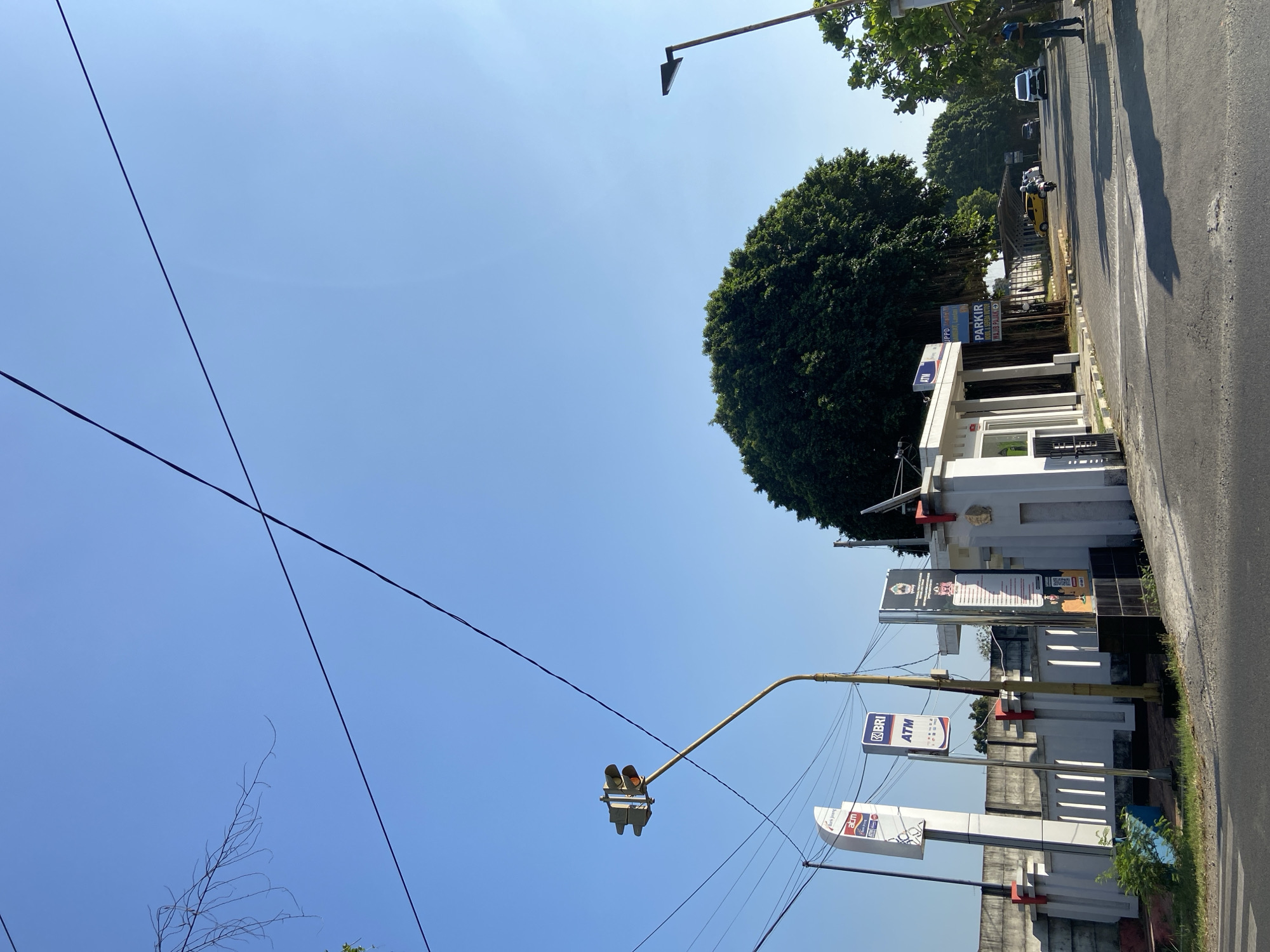 The image depicts a street scene in Margorejo, Indonesia, on a clear day with a bright blue sky. In the foreground, a paved road leads into the frame. To the left, a white wall with vertical slits runs along the side of the road, adorned with several signs, including ones for "ATM" and "BRI". A traffic light with yellow and red lights illuminated is positioned above the road, attached to a pole. Further down the road, a building with a white facade and a prominent entrance is visible. A large, dense green tree dominates the midground, its branches reaching towards the sky. Beyond the tree, more trees and foliage line the side of the road, with several parked cars and a motorcycle on a paved area. A street lamp stands to the right of the image. A person is standing on the right side of the road, near the edge of the frame. The overall scene suggests a typical urban or suburban environment with commercial and residential elements.