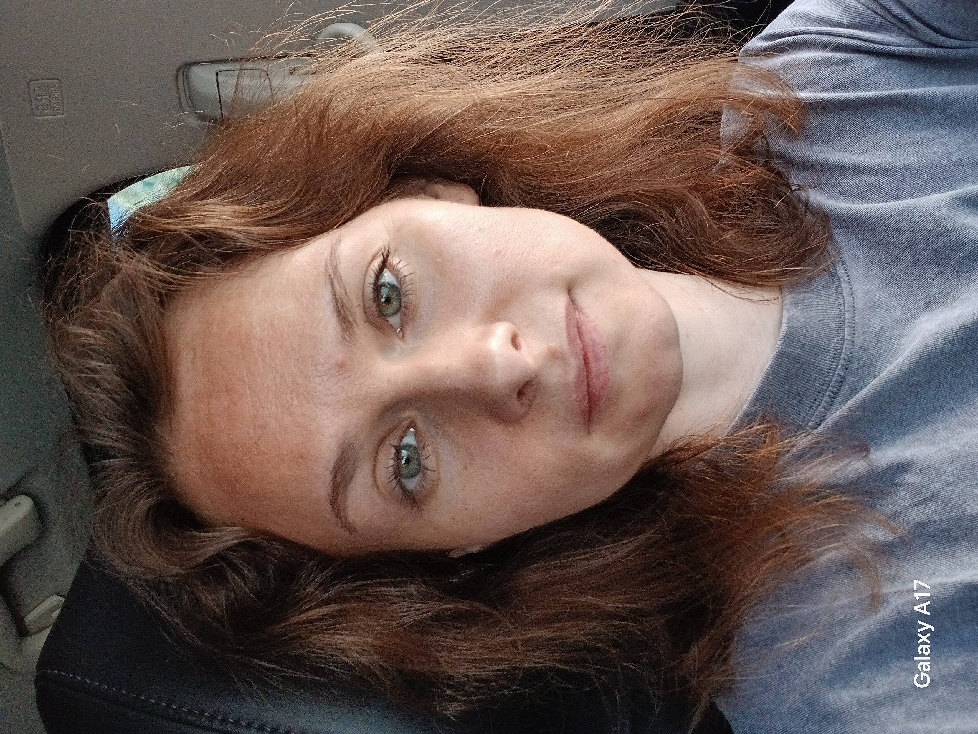 A young woman with fair skin and light auburn hair is lying down, likely in the back seat of a car. She is wearing a gray t-shirt. Her eyes are a light greenish-blue. The lighting suggests daylight. The background includes the car's interior, such as the headrest and ceiling. The lower right corner of the image has the watermark "Galaxy A17". There is no indication of Yekaterinburg or Russia.
